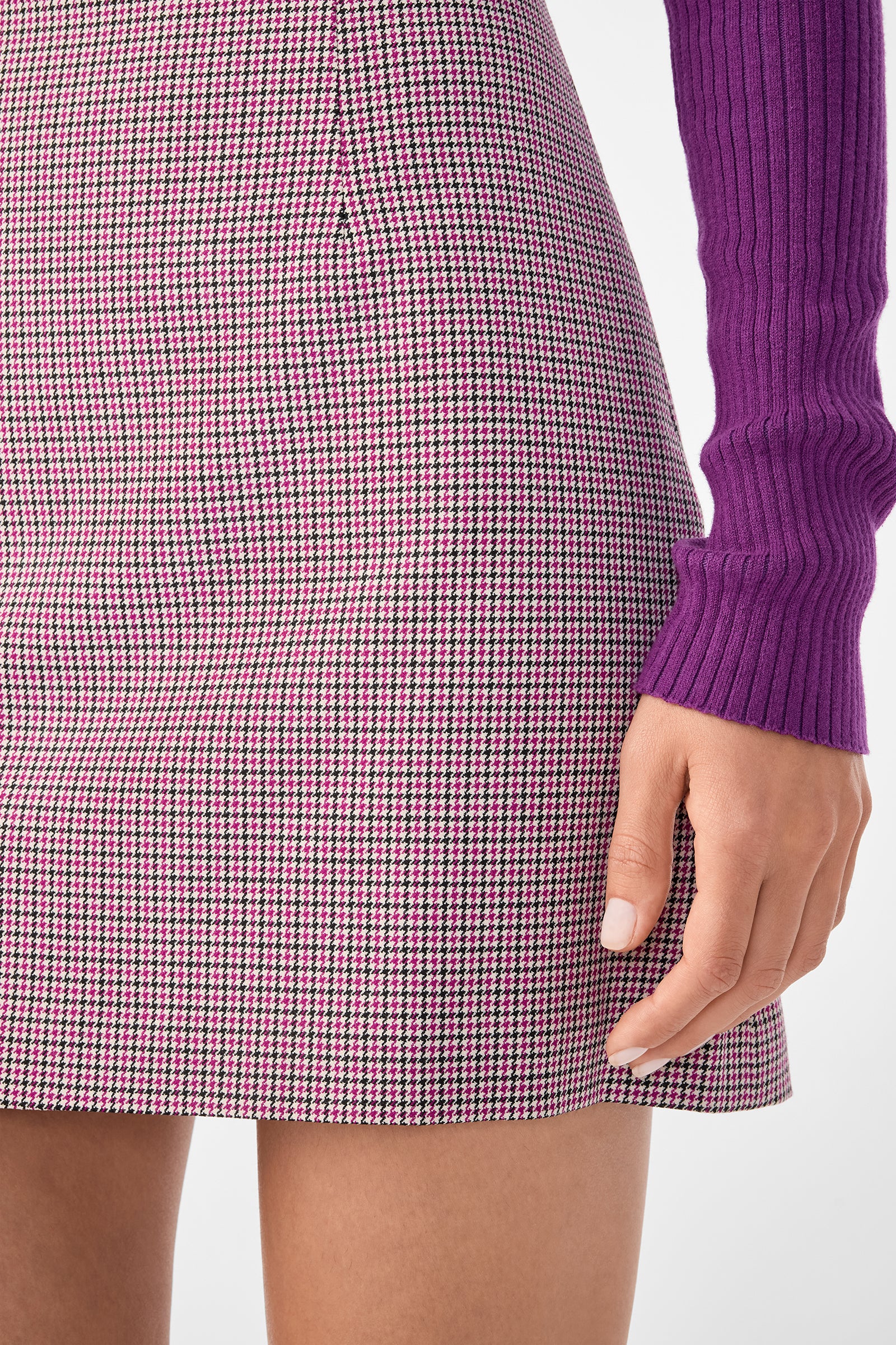 A close-up of a person wearing the High Rise Rico Skirt in pink and black houndstooth paired with a long-sleeve, ribbed purple top. Only the lower torso and right arm are visible.