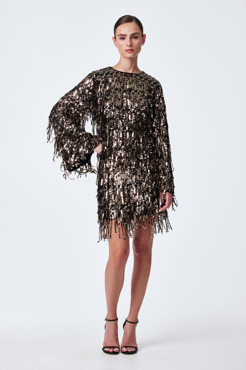 A woman wears the Rico Sequin Fringe Skirt, a black skirt adorned with gold and black sequins and fringe detailing. She pairs it with a top, has her hair pulled back, and wears black high-heeled sandals against a white background.