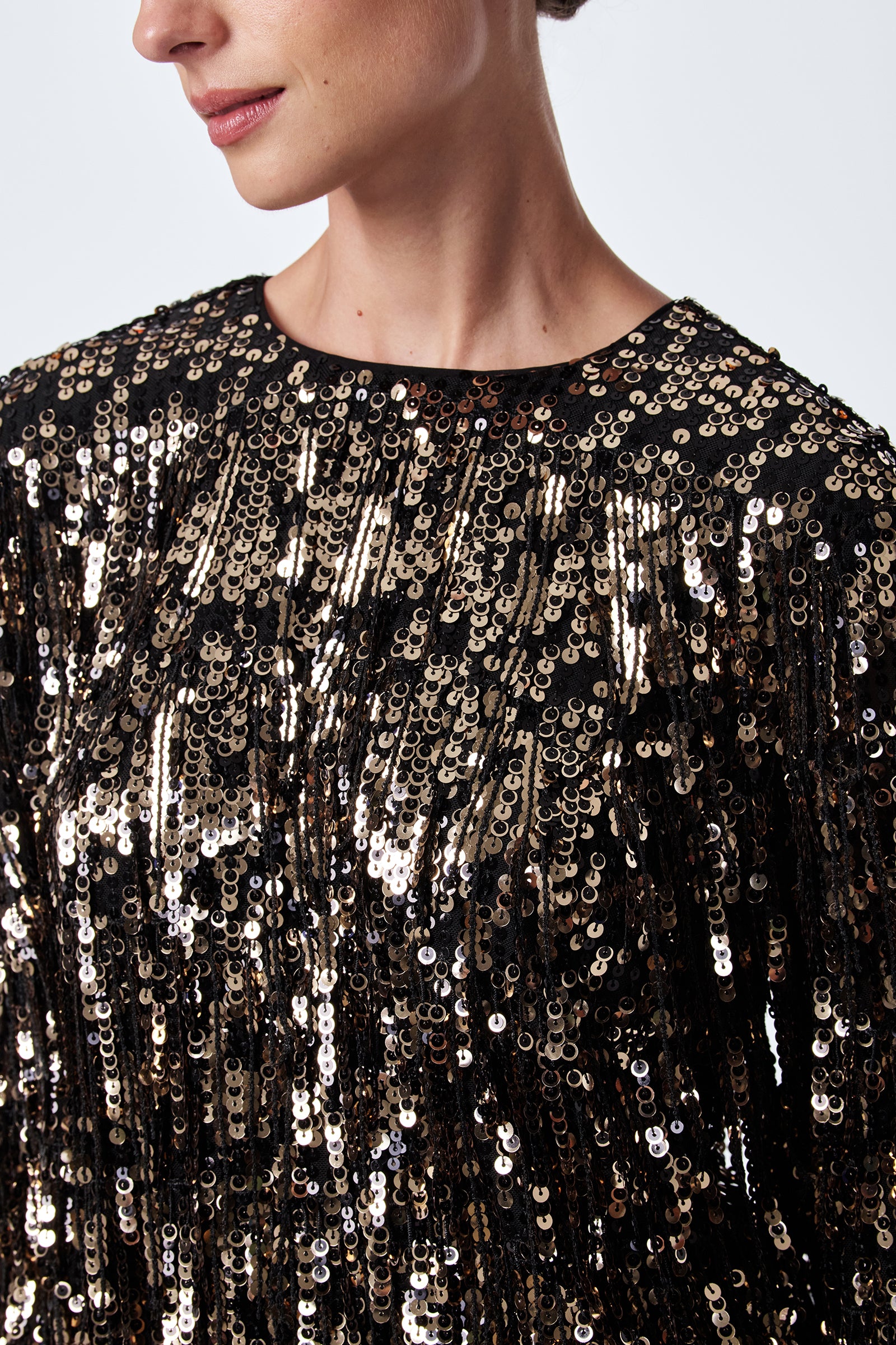 A person is shown from the shoulders up, wearing the Satine Sequin Fringe Top in black with shiny gold sequins. Their face is partially visible against a plain, light-colored background.