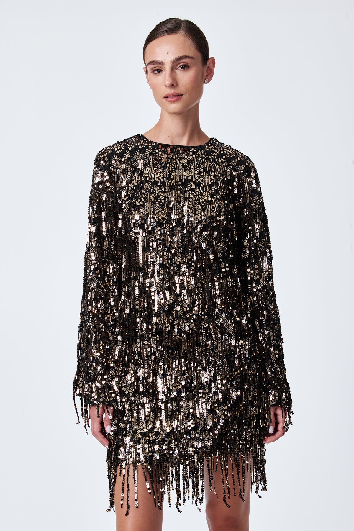 A woman stands against a plain background wearing the Satine Sequin Fringe Top, which features black and gold shimmering sequins with fringe detailing for a sparkling, textured effect.