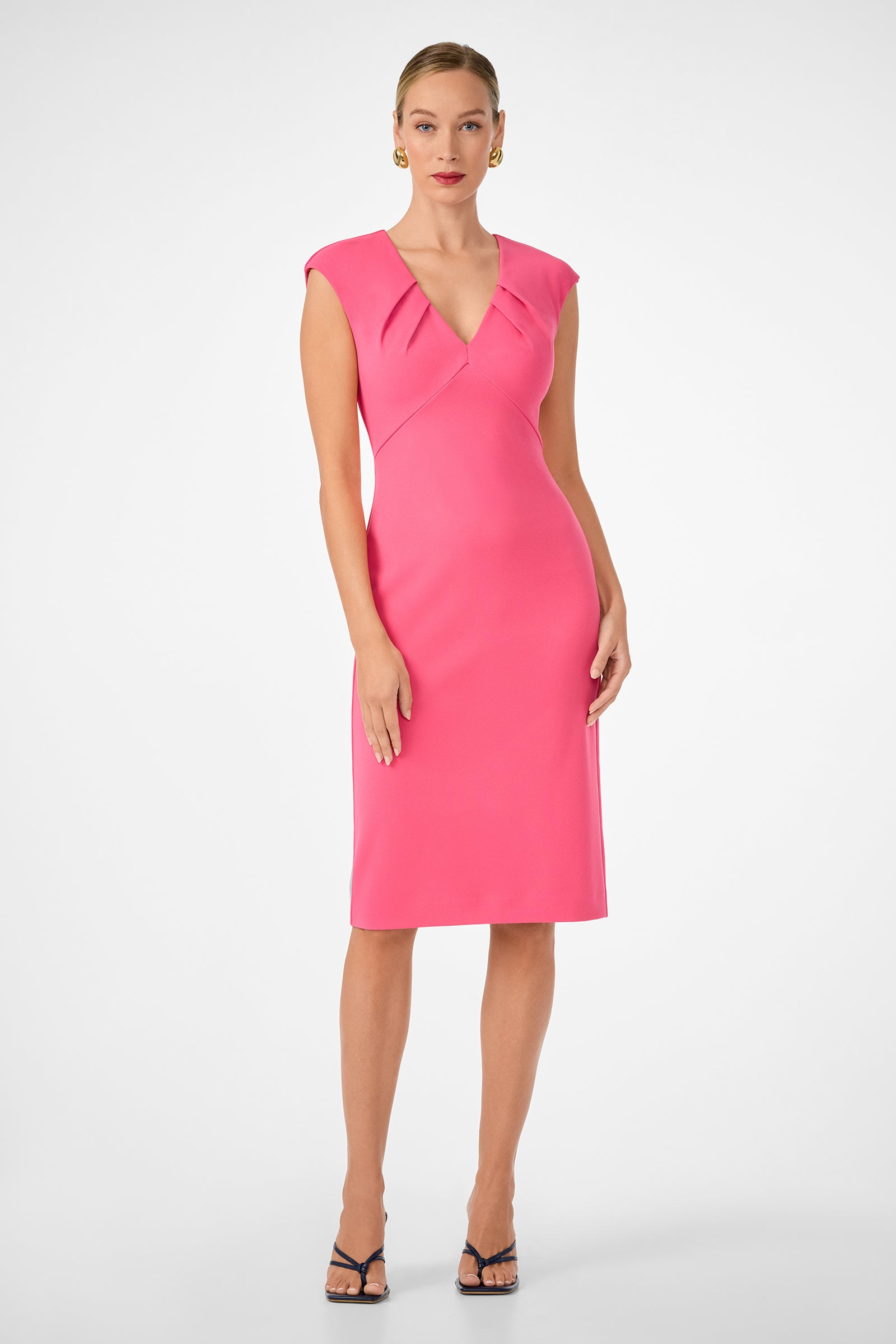 A woman wears the Vibrant Dress, a fitted knee-length bright pink dress with pleated shoulder details, V-neckline, and cap sleeves. She pairs it with gold earrings, red lipstick, and black heels against a plain white background.