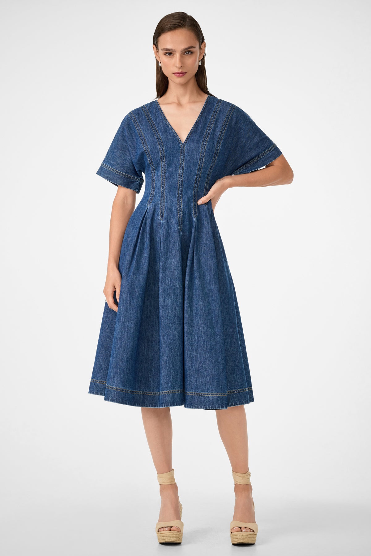A woman wears the Ashe 2 Short-Sleeve Denim Dress in indigo, featuring a knee-length flared skirt and V-neckline. She stands with one hand by her side and pairs the dress with beige platform sandals.