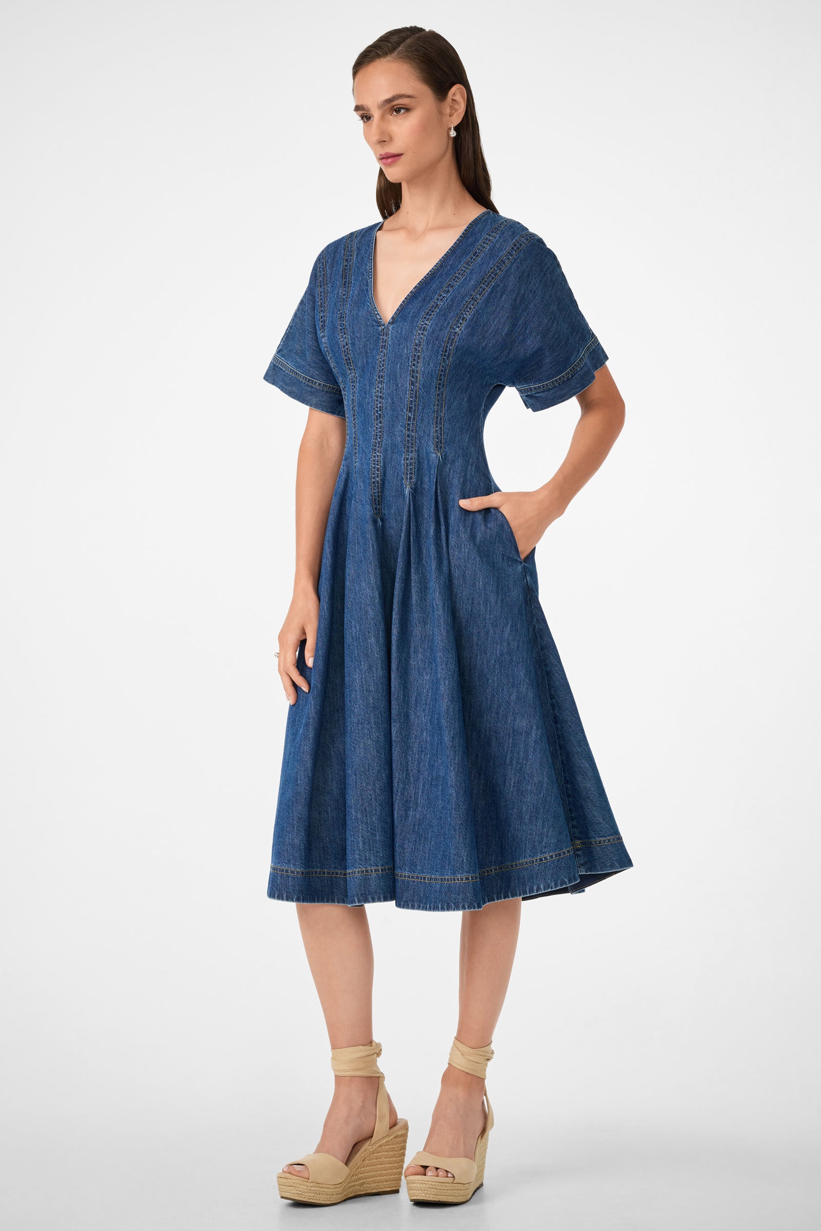 A woman models the Ashe 2 Short-Sleeve Denim Dress, a blue knee-length V-neck with short sleeves and pockets, paired with beige wedge sandals, against a plain white background.