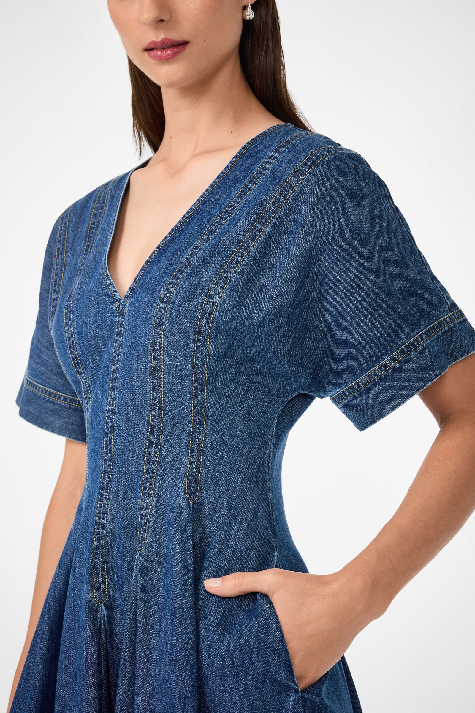 A woman models the Ashe 2 Short-Sleeve Denim Dress, featuring a V-neckline, short sleeves, and seam details. She poses with one hand in her pocket against a plain white background.