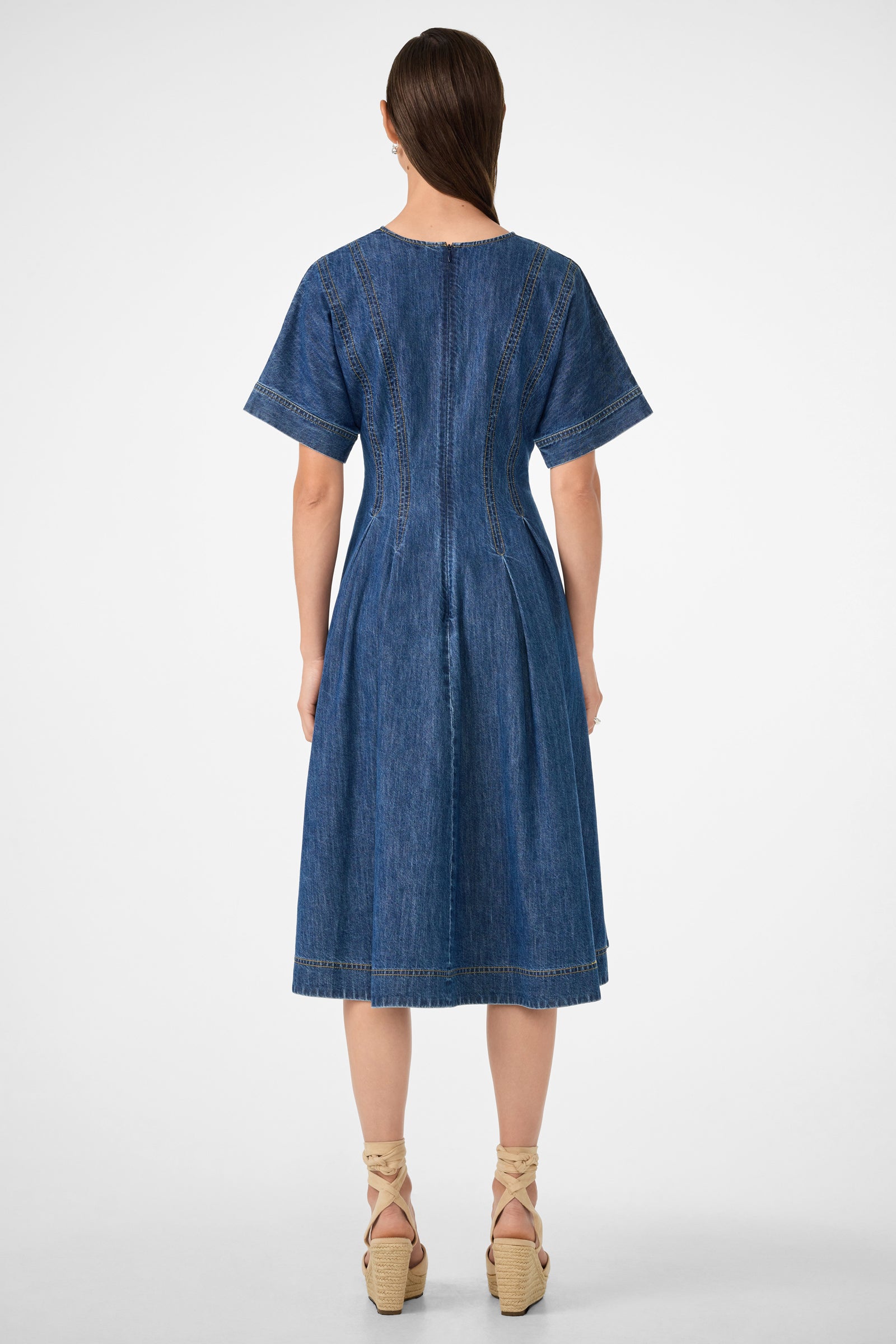A woman faces away, wearing the Ashe 2 Short-Sleeve Denim Dress—a blue denim midi with seam details. She pairs it with beige platform sandals and has straight brown hair pulled back.