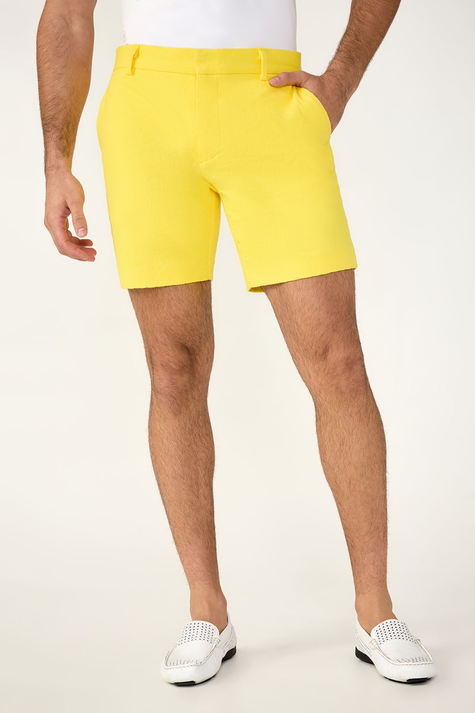 A person wears the Lawrence Short - Stretch Herringbone in Lavandou Lemon with a white tucked-in shirt and white loafers, hands in pockets. Only their lower torso and legs appear against a light background.