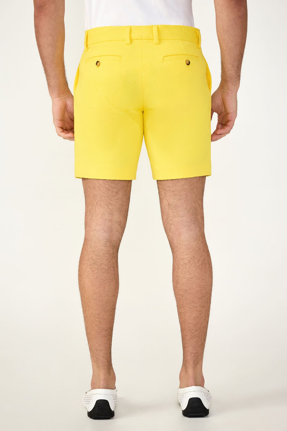A man stands facing away on a plain light background, wearing a white shirt, the Lawrence Short - Stretch Herringbone in bright yellow with belt loops and back pockets, and white slip-on shoes with black soles.