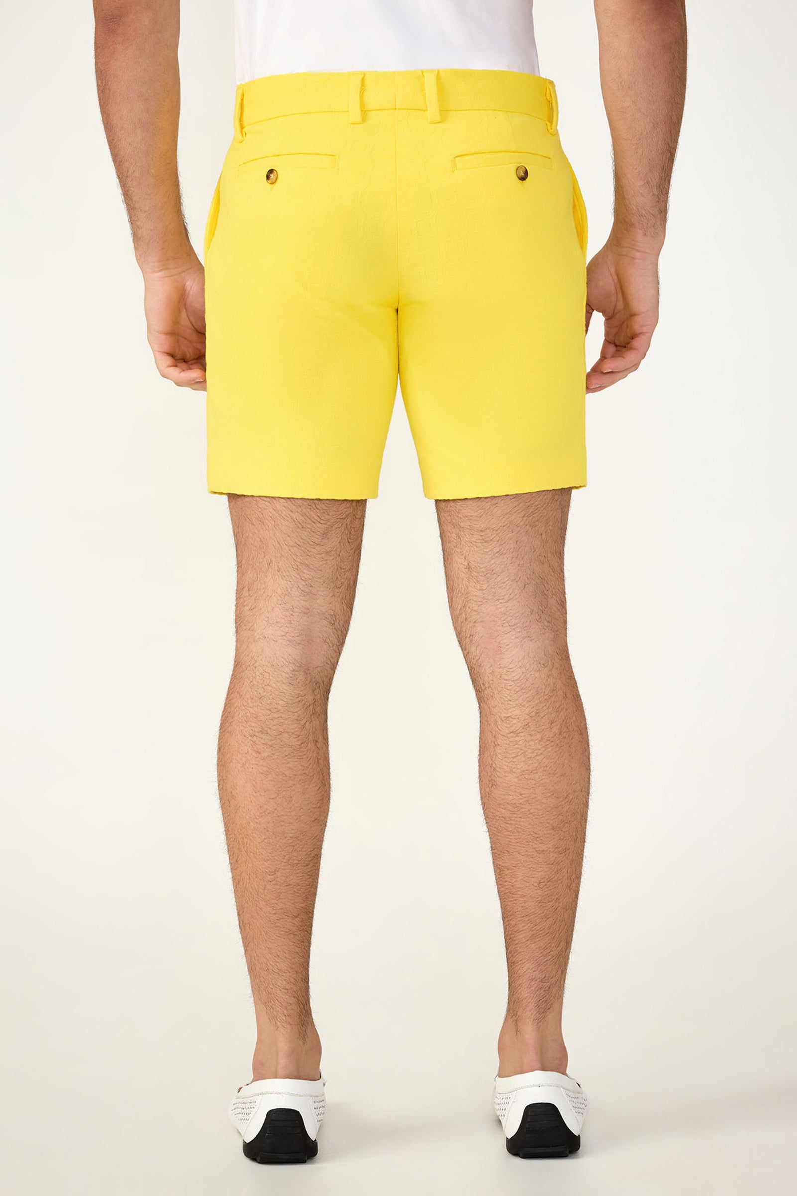 A man stands facing away on a plain light background, wearing a white shirt, the Lawrence Short - Stretch Herringbone in bright yellow with belt loops and back pockets, and white slip-on shoes with black soles.