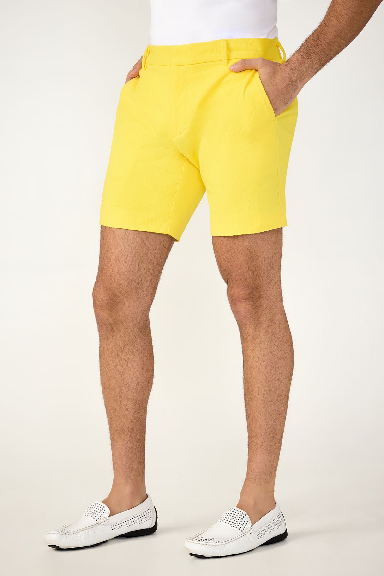 Someone wearing the Lawrence Short - Stretch Herringbone in bright yellow, paired with white loafers, stands with hands in pockets against a plain light background. Only the lower half of the body is shown.