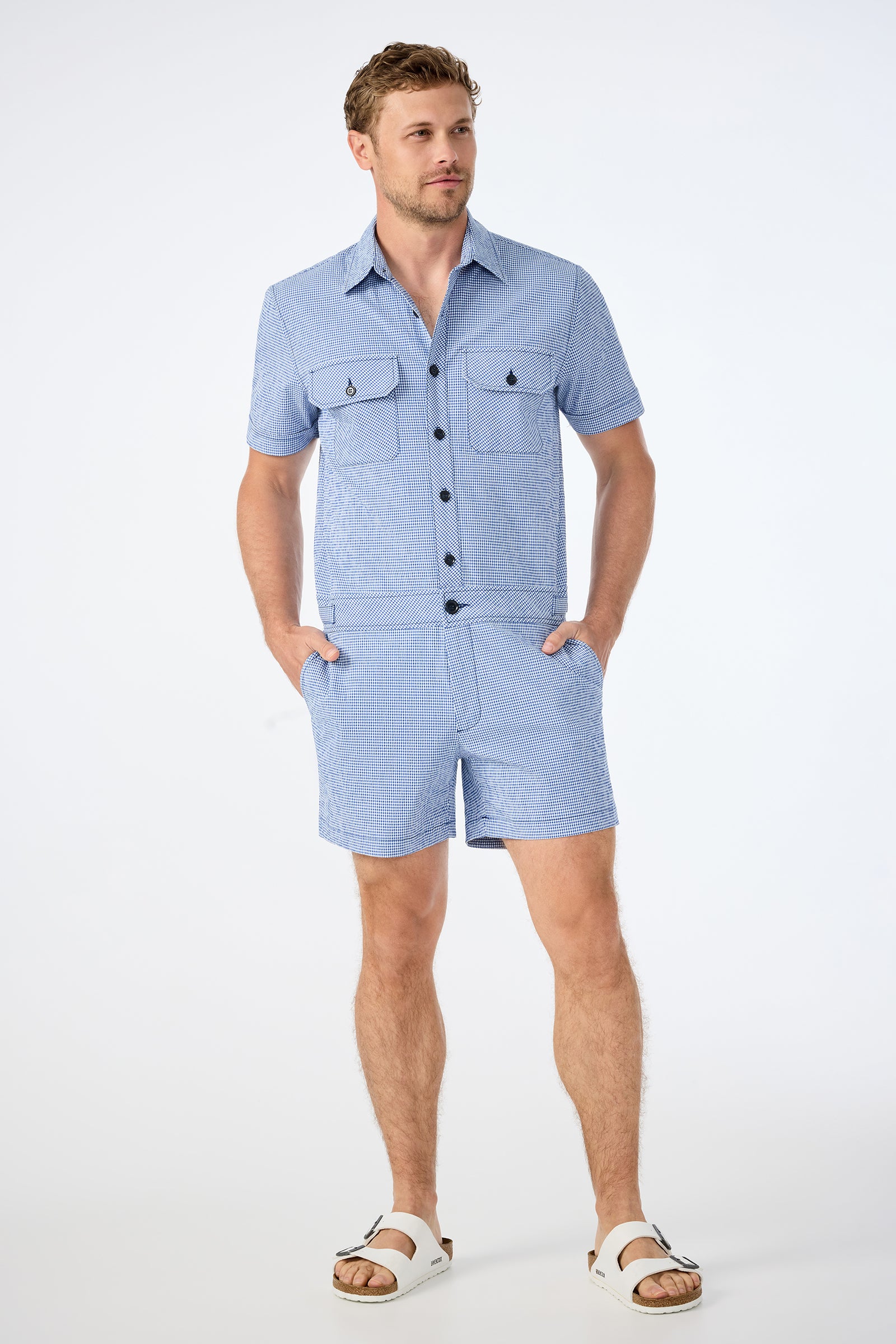 A man with short brown hair and a beard stands against a plain background wearing the Sherman Short Jumpsuit in Ocean Gingham with front pockets and white sandals, his hands in his pockets.