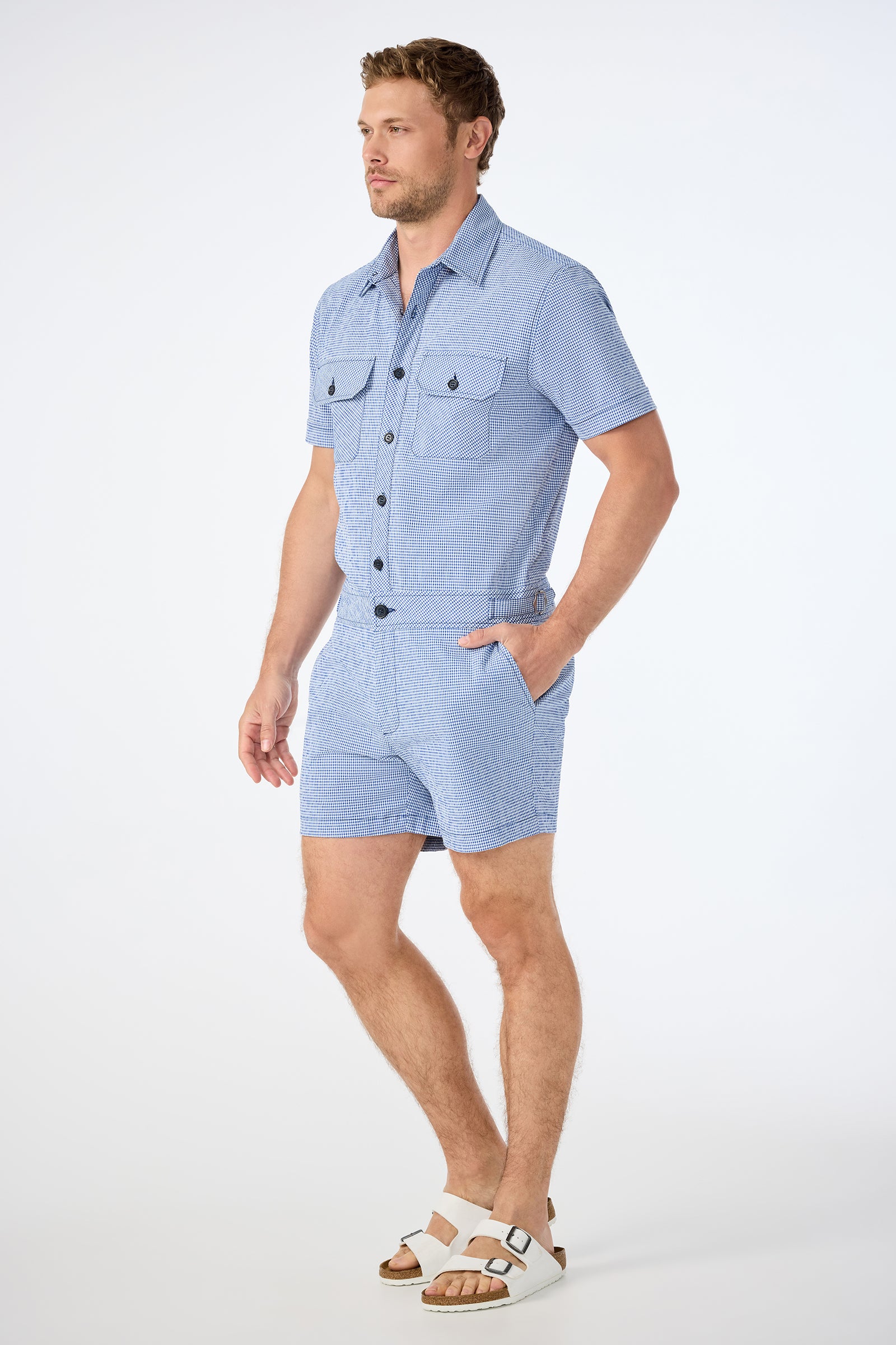 A man stands against a plain background wearing the Sherman Short Jumpsuit in Ocean Gingham with white sandals. His left hand is in his pocket as he looks off to the side.