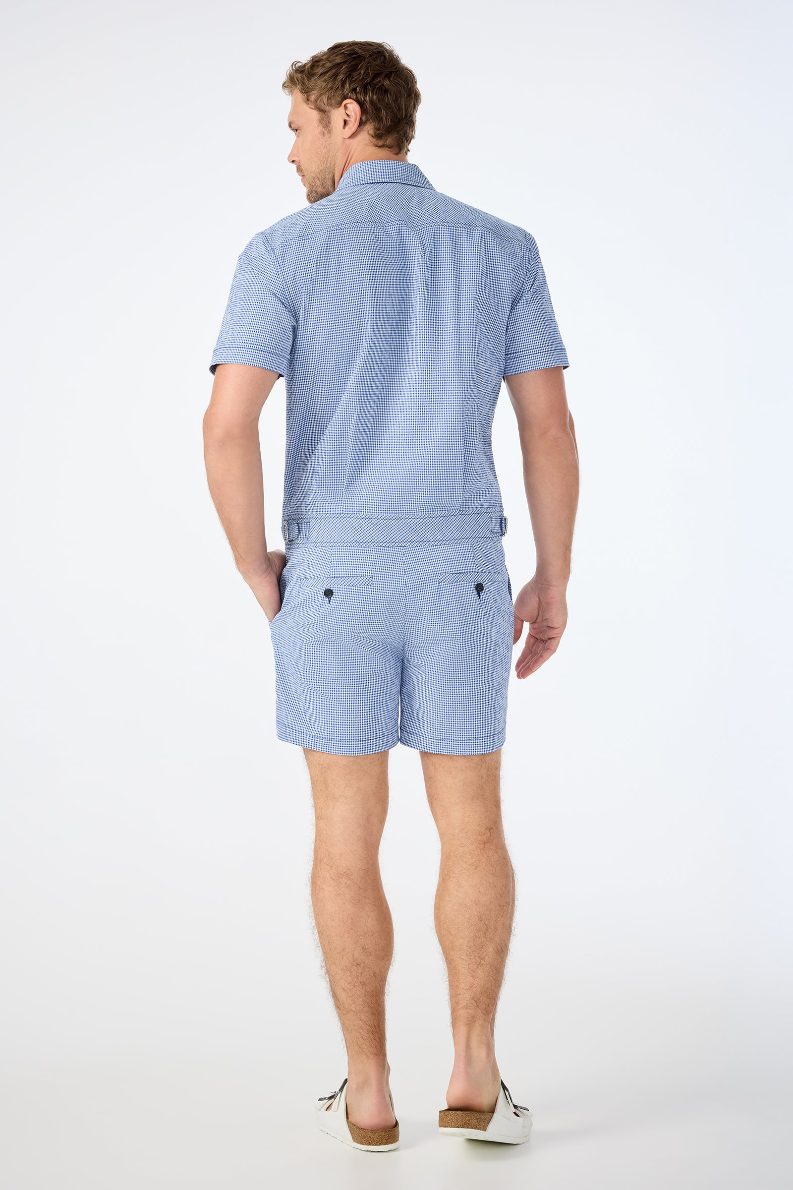 A man faces away from the camera, wearing the Sherman Short Jumpsuit in Ocean Gingham with white slip-on shoes against a plain white background.