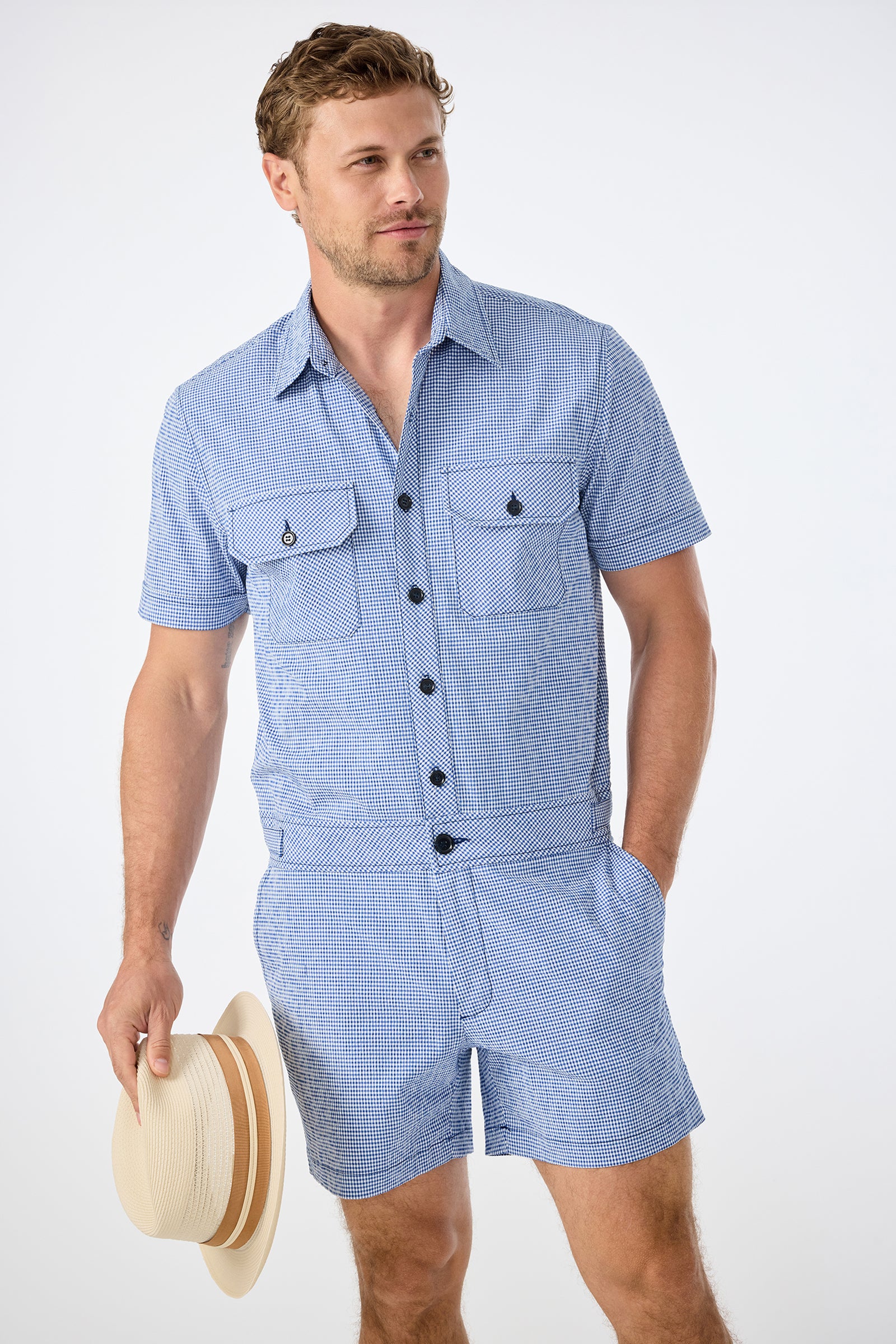 A man wearing the Sherman Short Jumpsuit in Ocean Gingham holds a beige straw hat with a tan band in his right hand, standing against a plain white background.