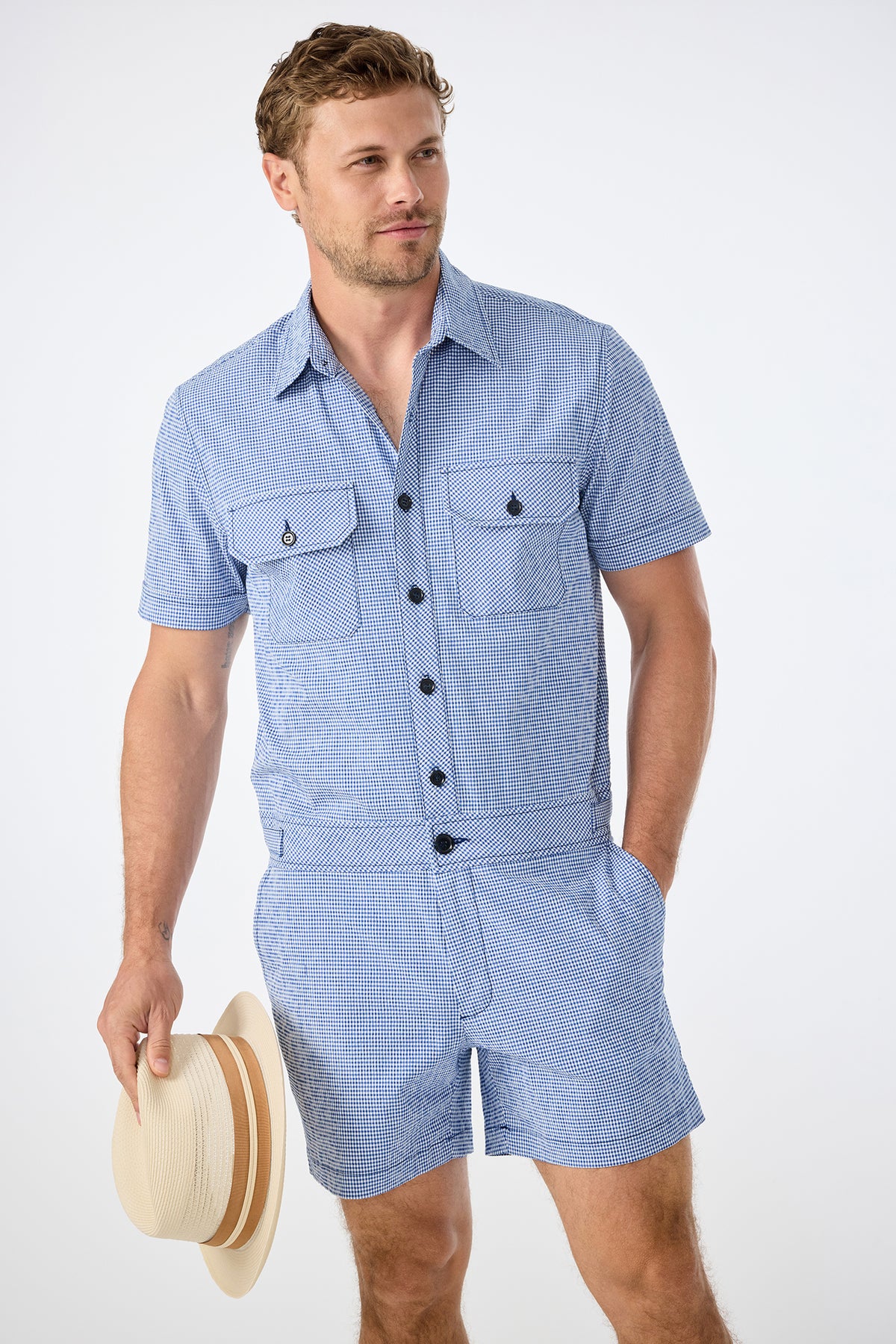 A man wearing the Sherman Short Jumpsuit in Ocean Gingham holds a beige straw hat with a tan band in his right hand, standing against a plain white background.