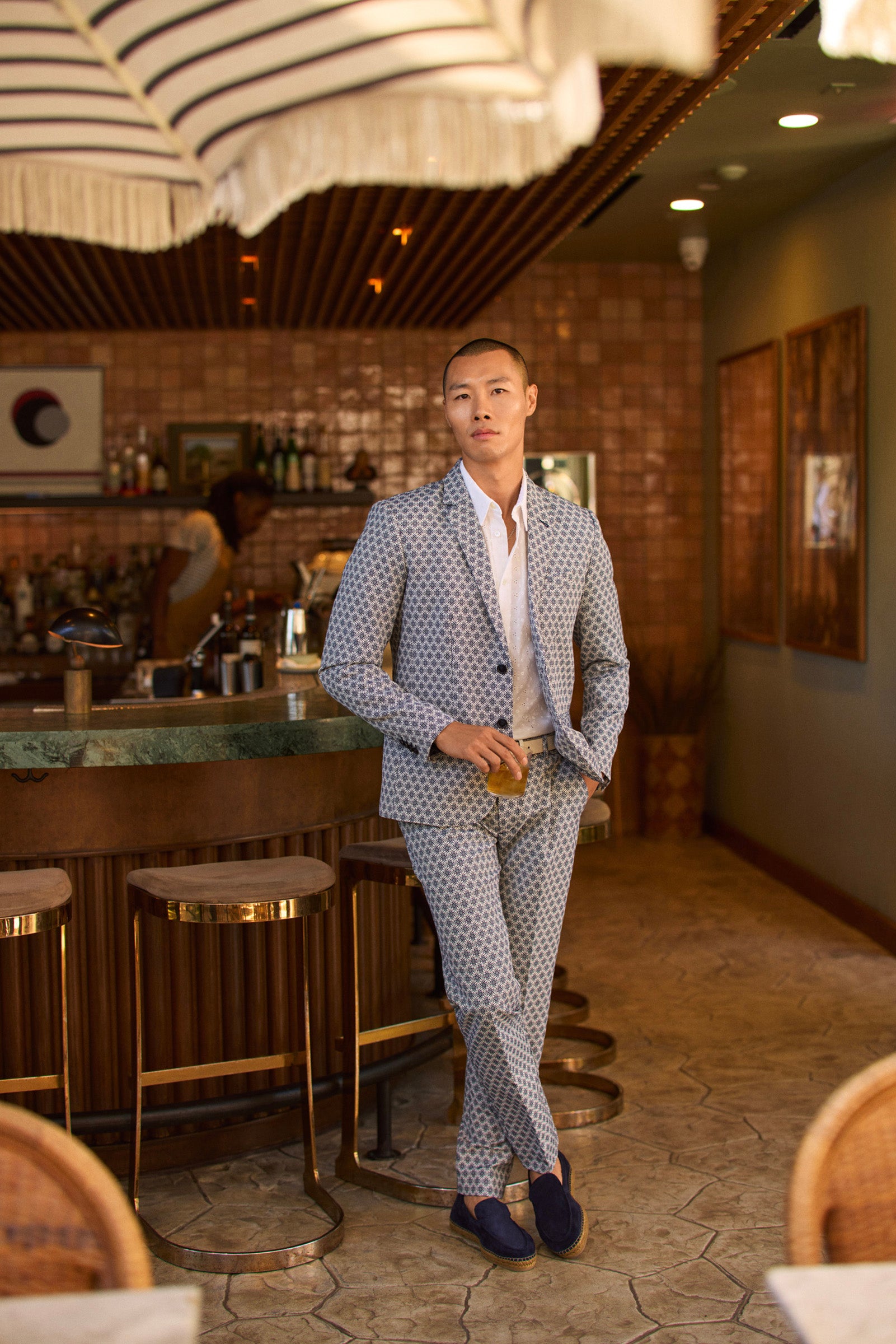 Wearing the Clyde Slim Trouser - Daisy Jacquard and blue loafers, a person stands by a warmly lit bar with tiled walls and stools, holding a drink as someone works behind the counter.