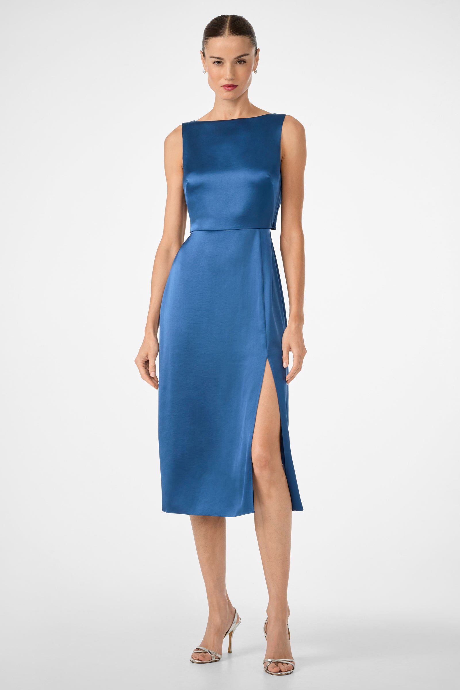 A woman wears the Mochi Sleeveless Midi Dress in Sumi Blue, featuring a fitted silhouette and high side slit, styled with silver high-heeled sandals. She stands against a plain white background.