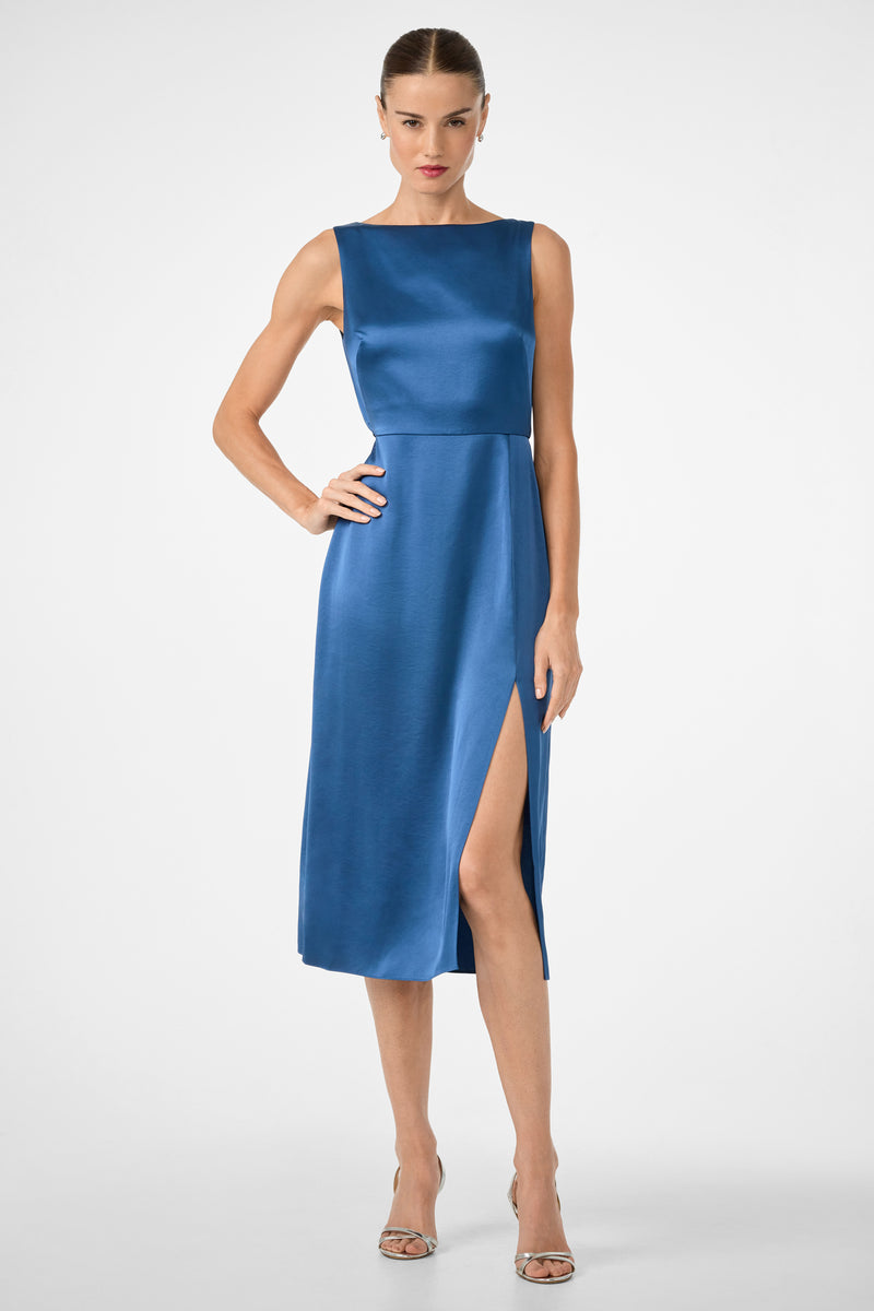A woman poses against a plain background in the Mochi Sleeveless Midi Dress, a blue satin, knee-length dress featuring a high side slit. She pairs it with silver heels and slicked-back hair, with one hand on her hip.
