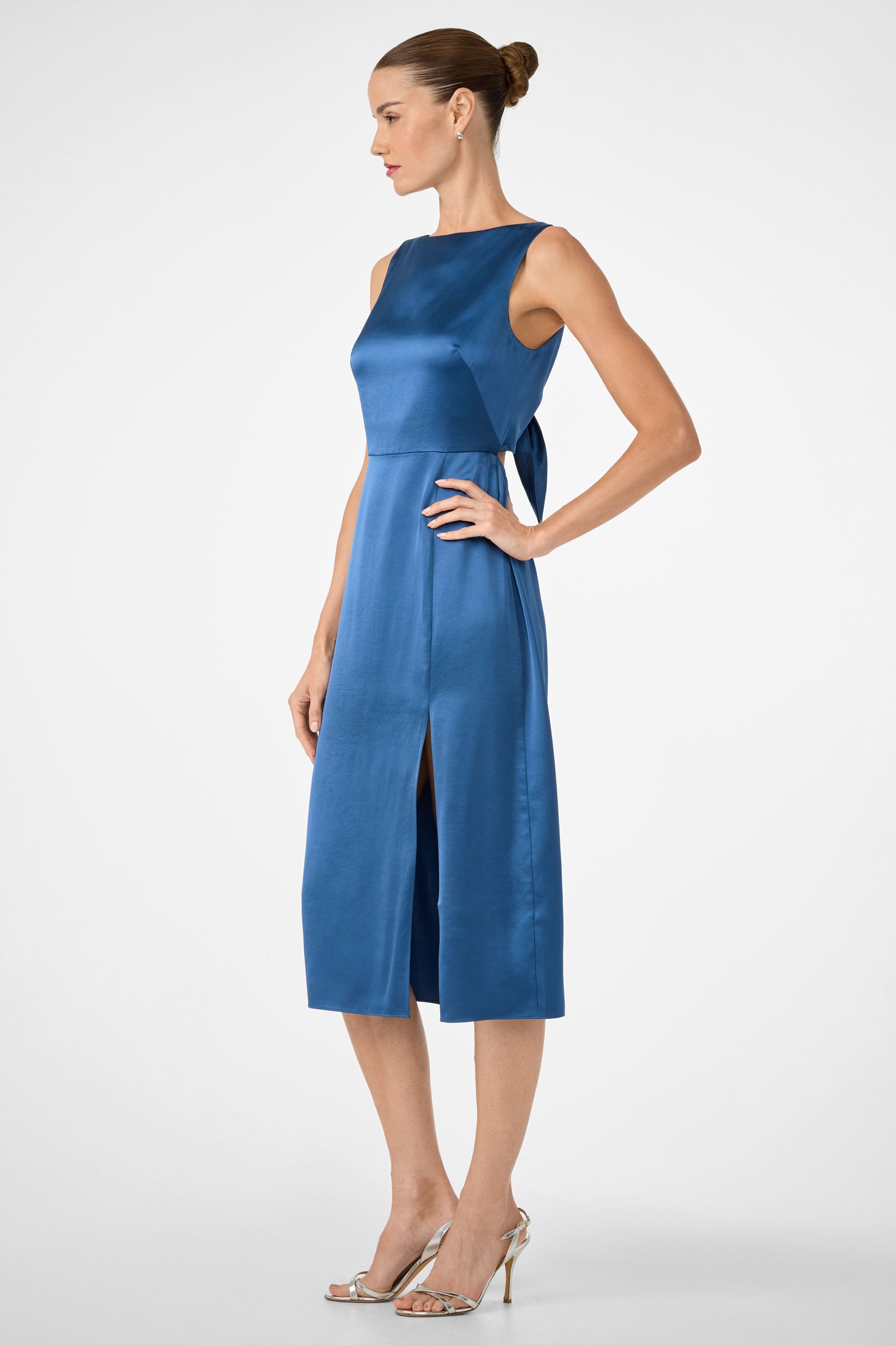 Woman in the Mochi Sleeveless Midi Dress—blue satin, knee-length, sleeveless with side slit and tie-back detail—standing in side profile with hair in a bun, wearing silver high-heeled sandals against a plain background.