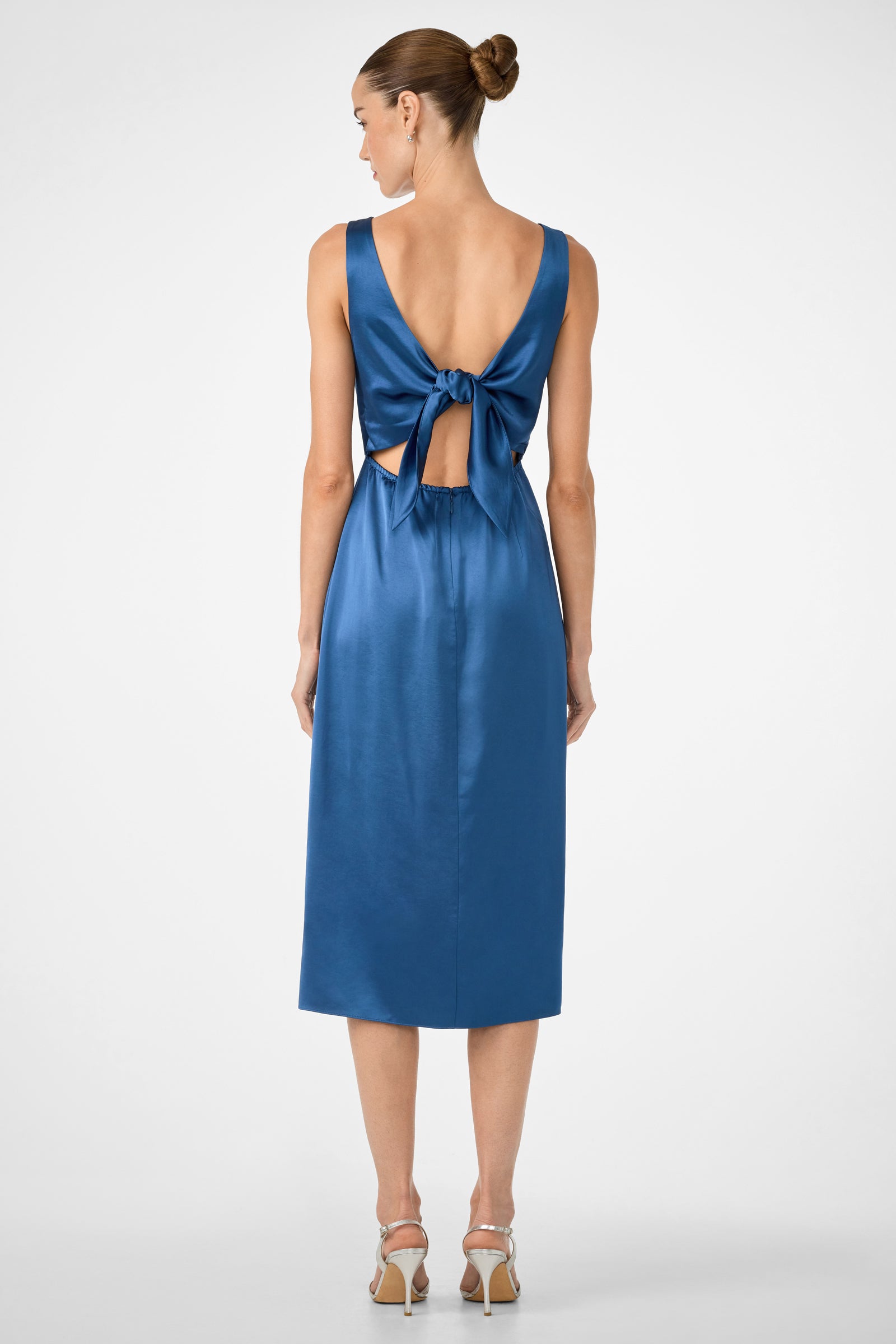 A woman with her hair in a neat bun wears the Mochi Sleeveless Midi Dress, featuring a shiny blue fabric, low back, large waist bow, and midi length. She pairs it with strappy high-heeled shoes and faces away from the camera.