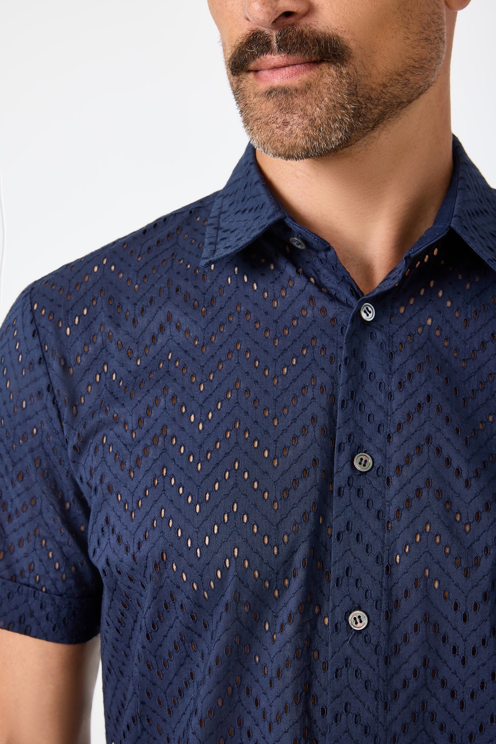A man with a mustache wears the Branson Shirt - Cotton Eyelet, a semi-sheer, short-sleeve navy button-up featuring a chevron eyelet pattern. The shirt reveals glimpses of skin; only his chest and lower face are visible.