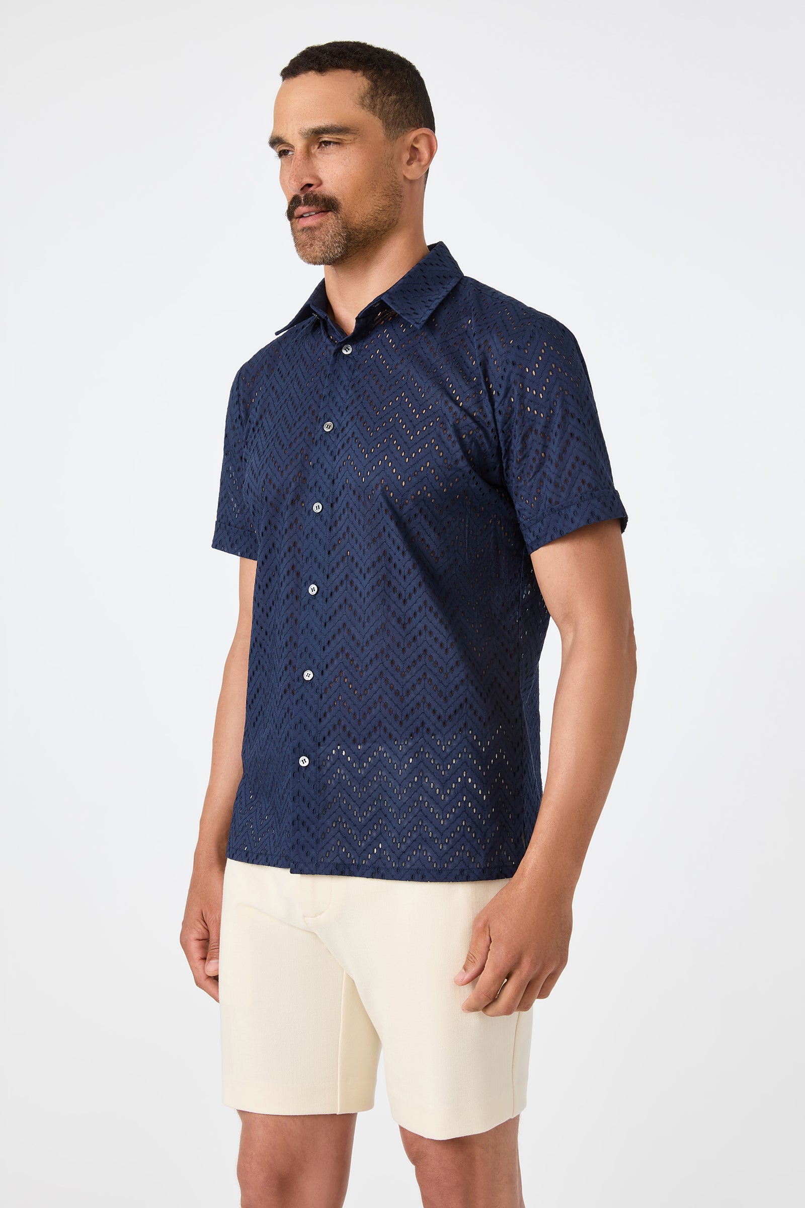 A man wears the Branson Shirt - Cotton Eyelet, a navy blue short-sleeve button-up with a zigzag eyelet pattern, paired with beige shorts, against a plain white background.