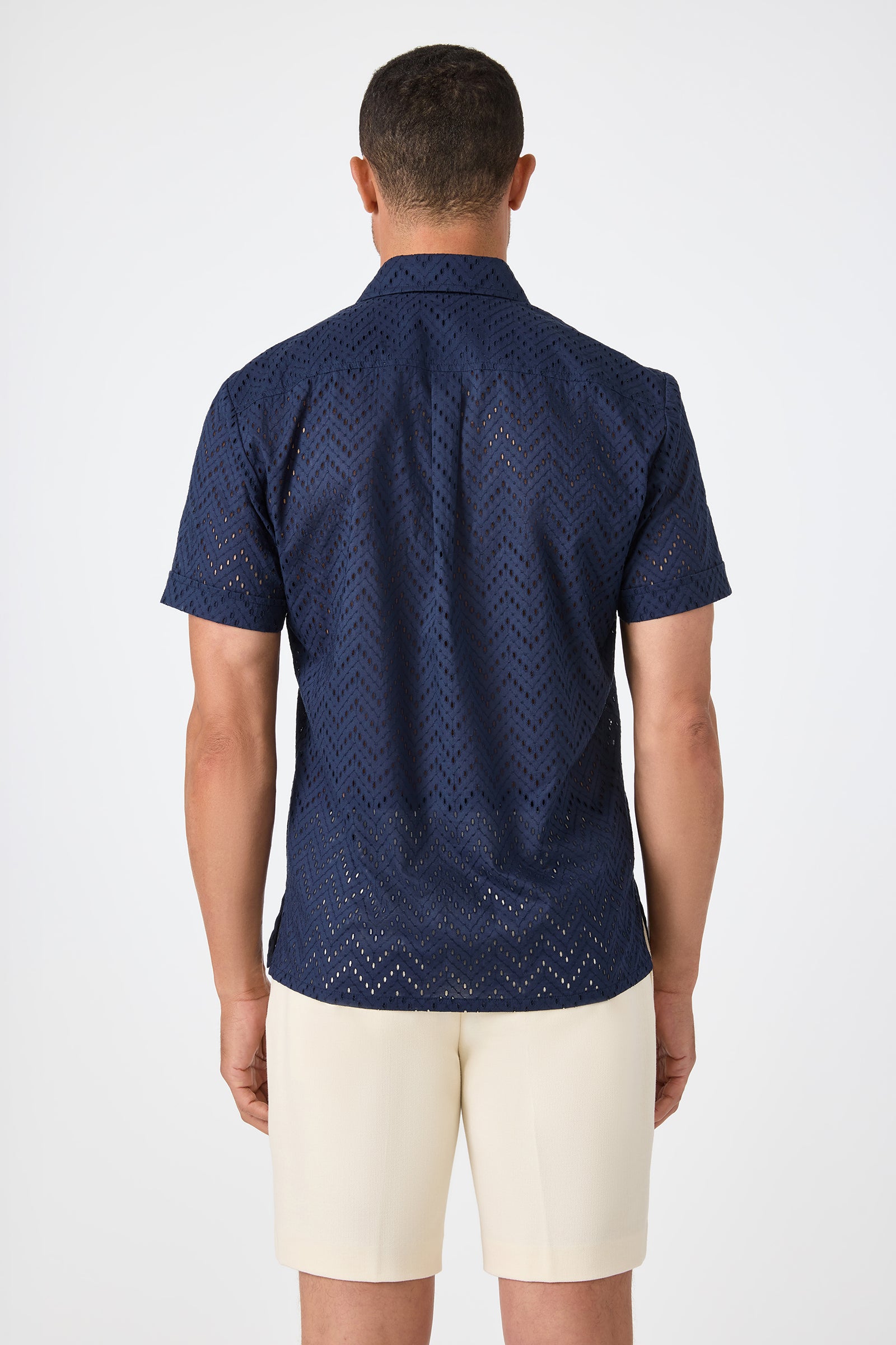 A man faces away, wearing the Branson Shirt - Cotton Eyelet in navy with cream-colored shorts, set against a plain white background.