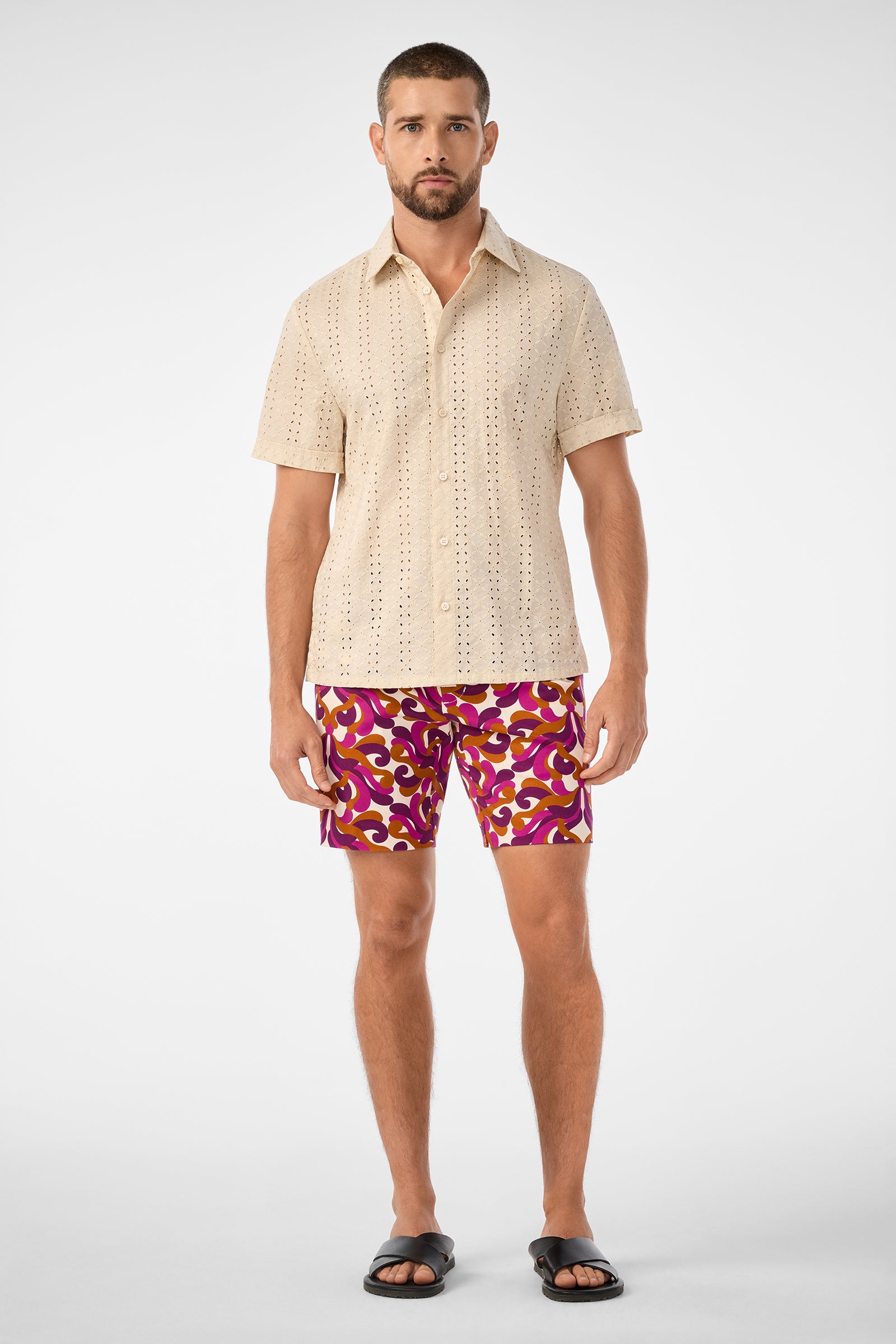 A man with short hair and a trimmed beard models the Branson Shirt—a beige short-sleeve button-up with eyelet details—pairing it with patterned pink and yellow shorts and black sandals against a plain background.