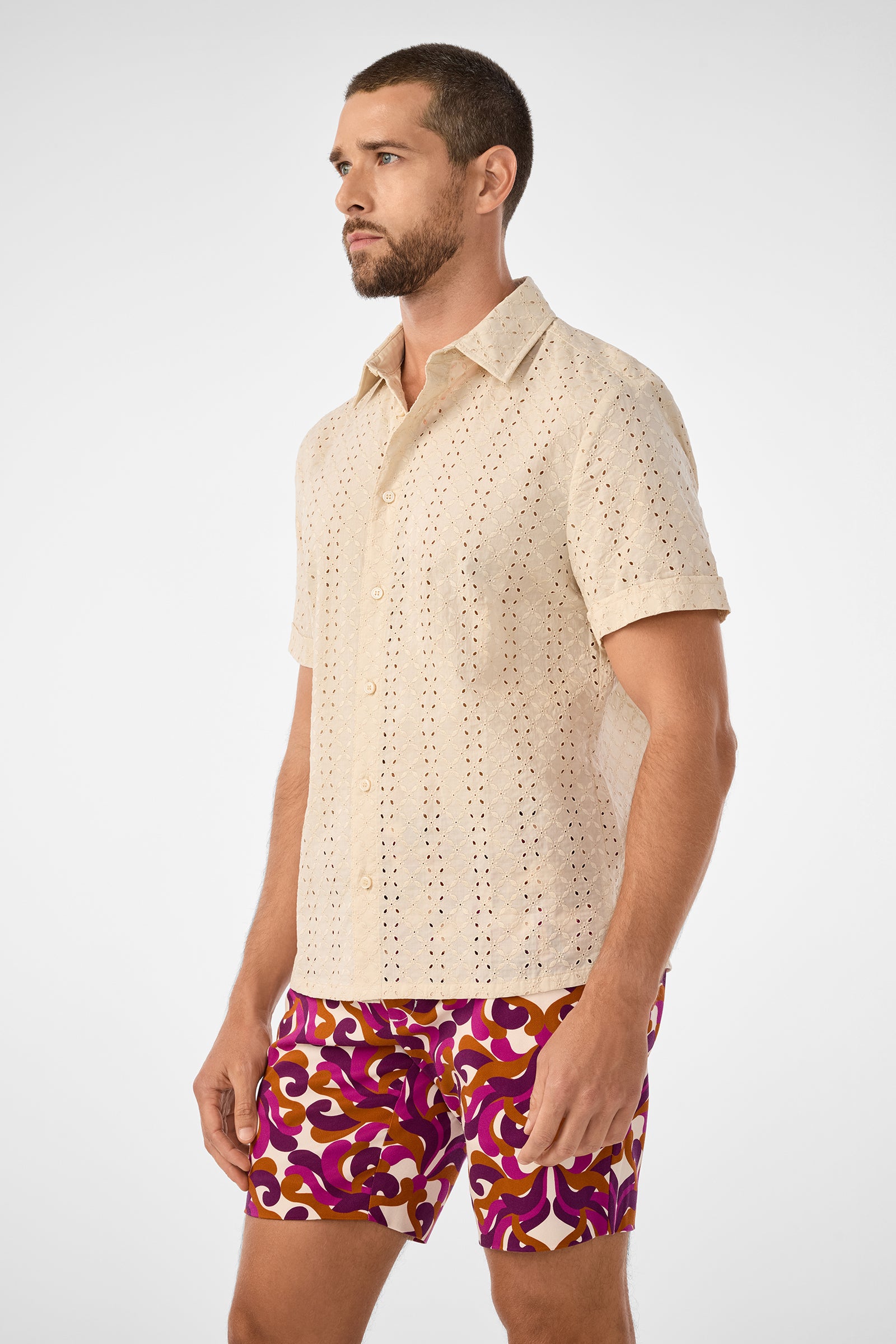 A man with short hair and a beard wears the beige Branson Shirt with eyelet details and colorful patterned shorts. He stands facing slightly left against a plain white background.