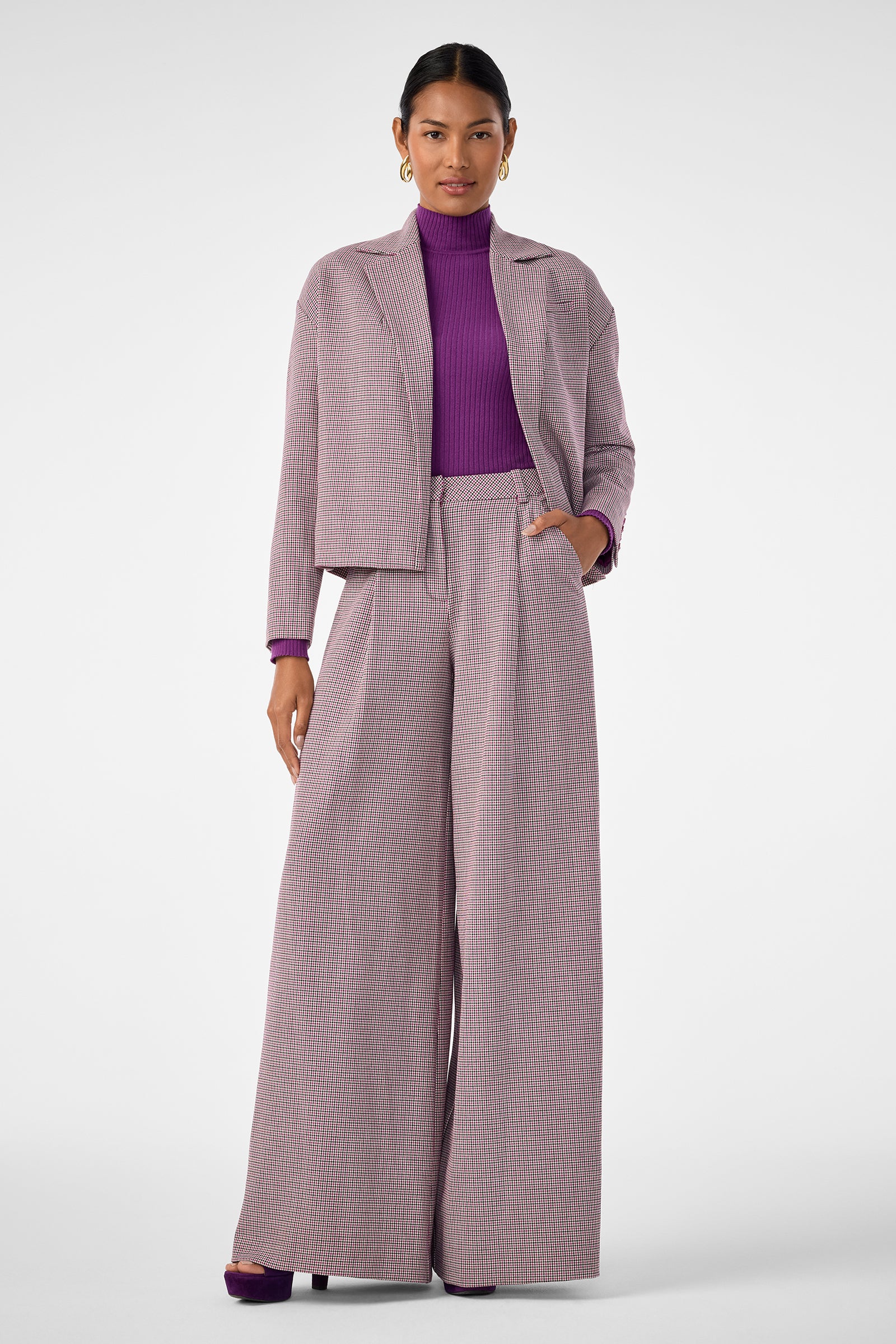 A woman stands against a white background wearing the Solani Pant, a light gray and pink checked wide-leg suit with a matching jacket, styled with a purple turtleneck top and purple heels.