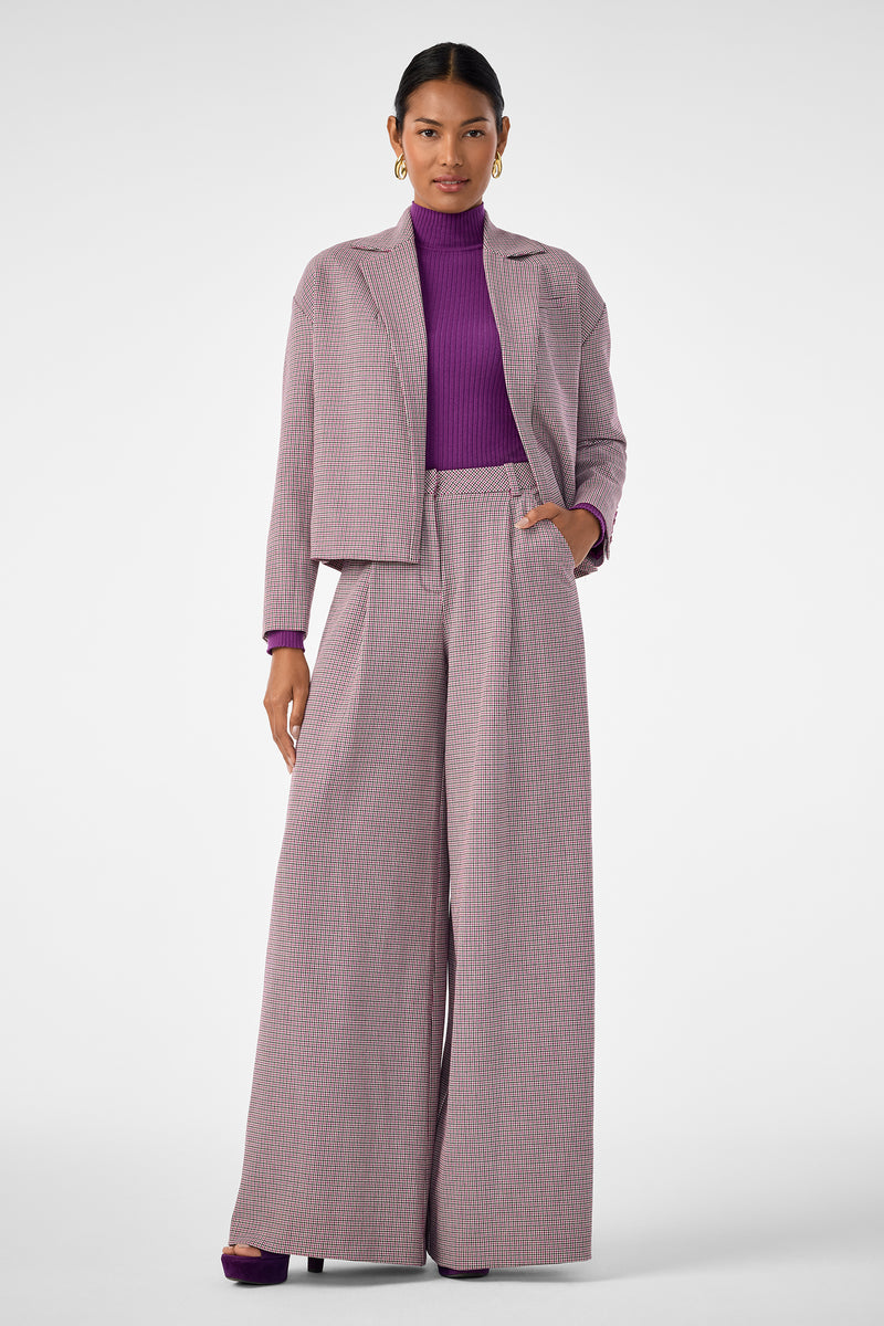A woman stands against a white background wearing the Solani Pant, a light gray and pink checked wide-leg suit with a matching jacket, styled with a purple turtleneck top and purple heels.