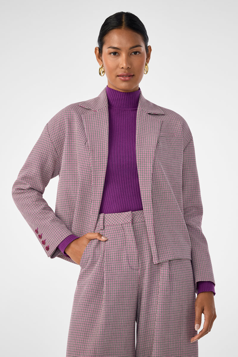 A woman with dark hair in a low bun wears gold hoops, a purple ribbed turtleneck, wide-leg plaid trousers, and the Ayla Jacket—a relaxed blazer—while standing against a light gray background.