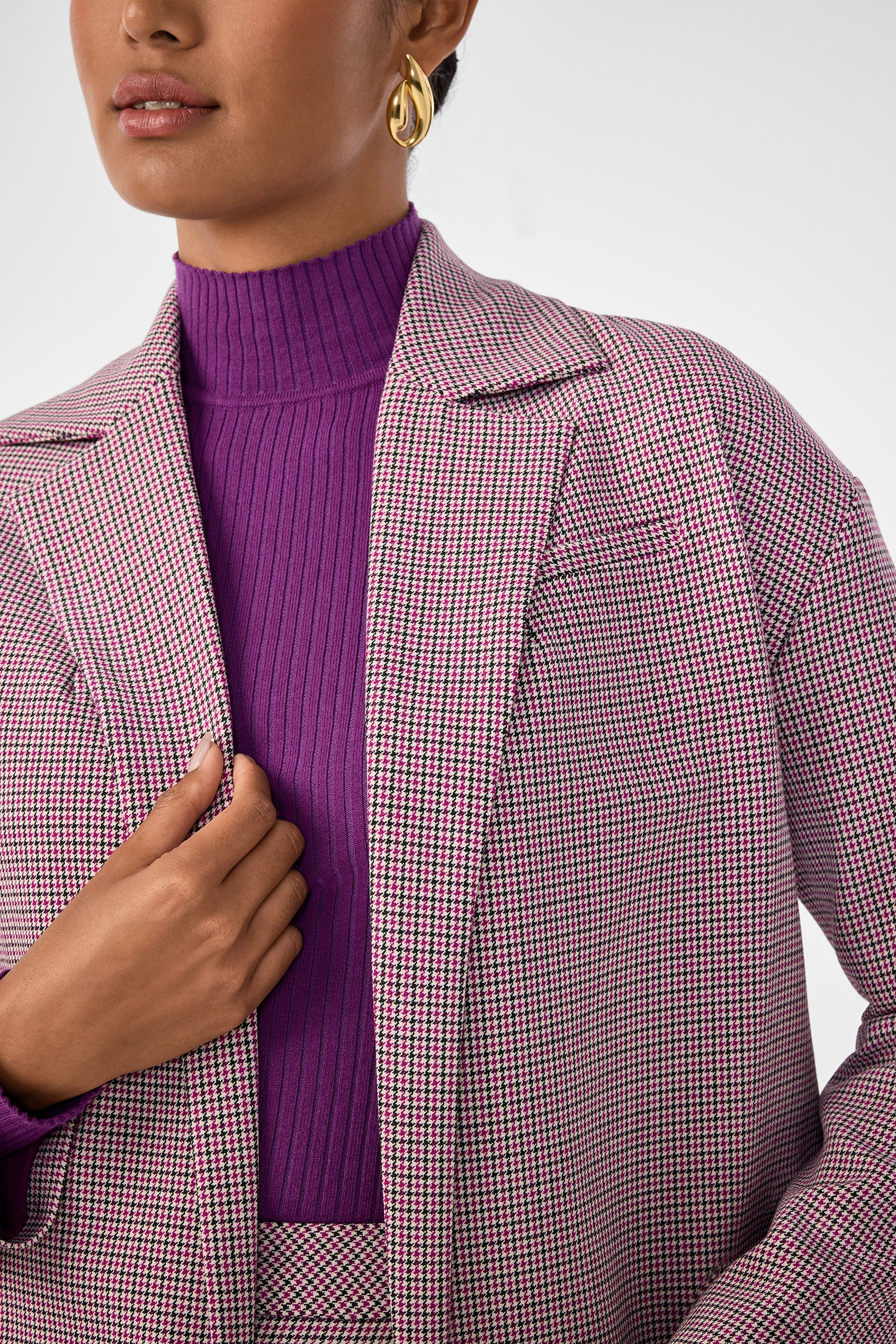 A person wears the Ayla Jacket, a pink and black houndstooth blazer over a purple ribbed turtleneck, accessorized with gold hoop earrings. Only their lower face and upper body are shown as they hold the blazer with one hand.