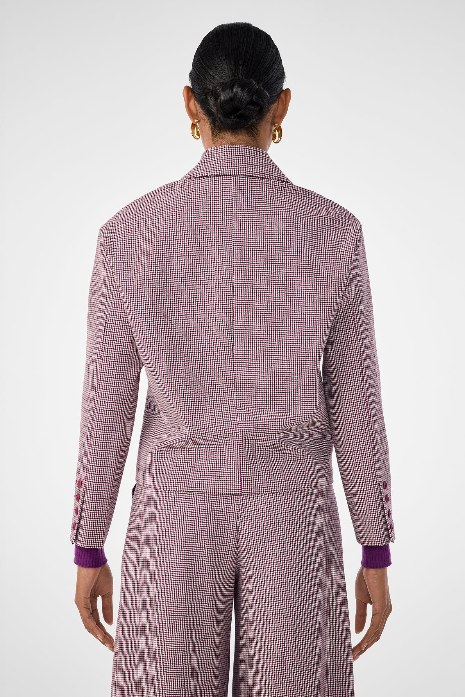 A person with dark hair in a neat bun is shown from behind, wearing the pink and white checkered Ayla Jacket with matching wide-leg pants and gold hoop earrings against a plain, light-colored background.