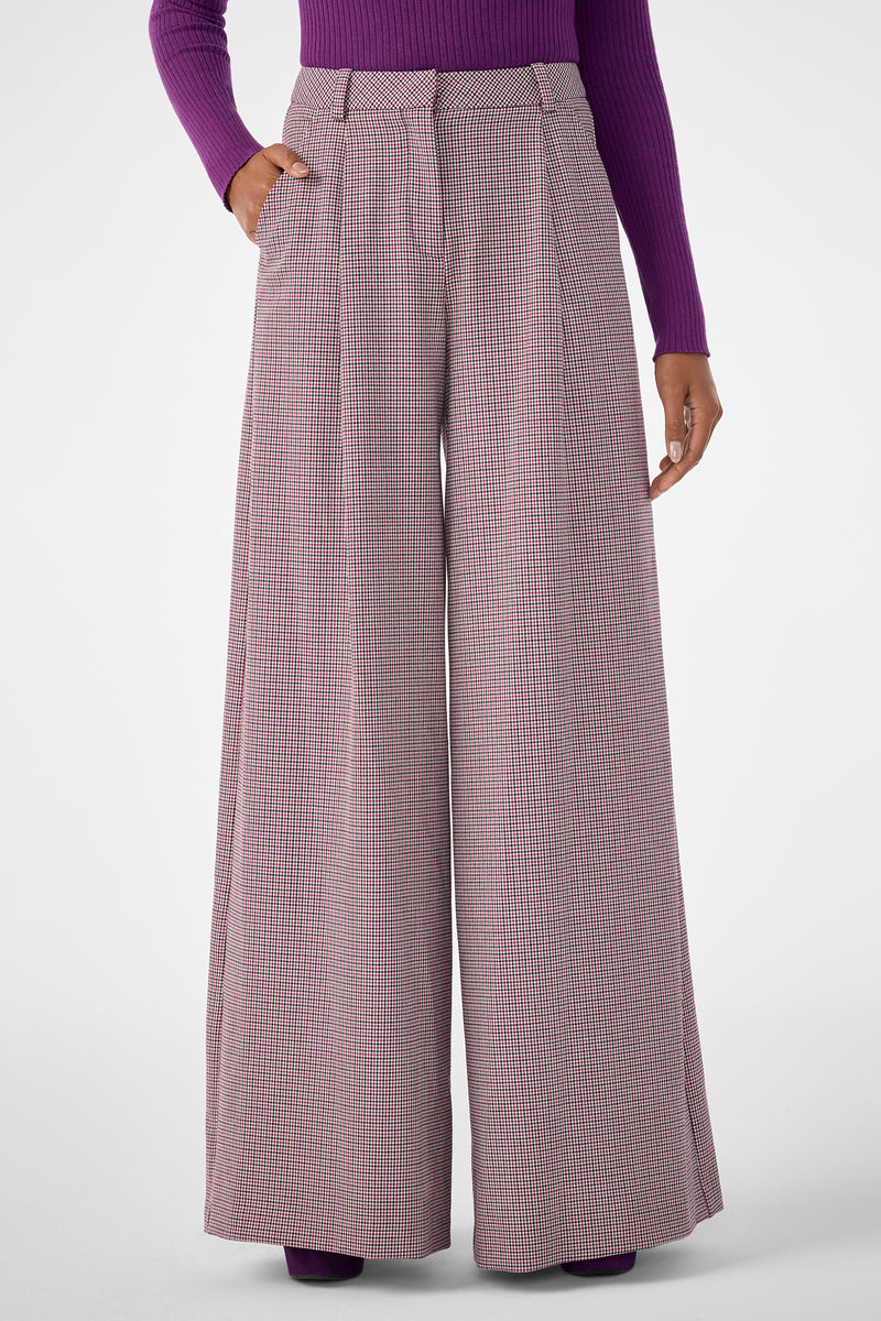 A person wears the Solani Pant, featuring high-waisted, wide-leg plaid in pink and grey, paired with a purple ribbed long-sleeve top. One hand is in their pocket, standing against a plain background.