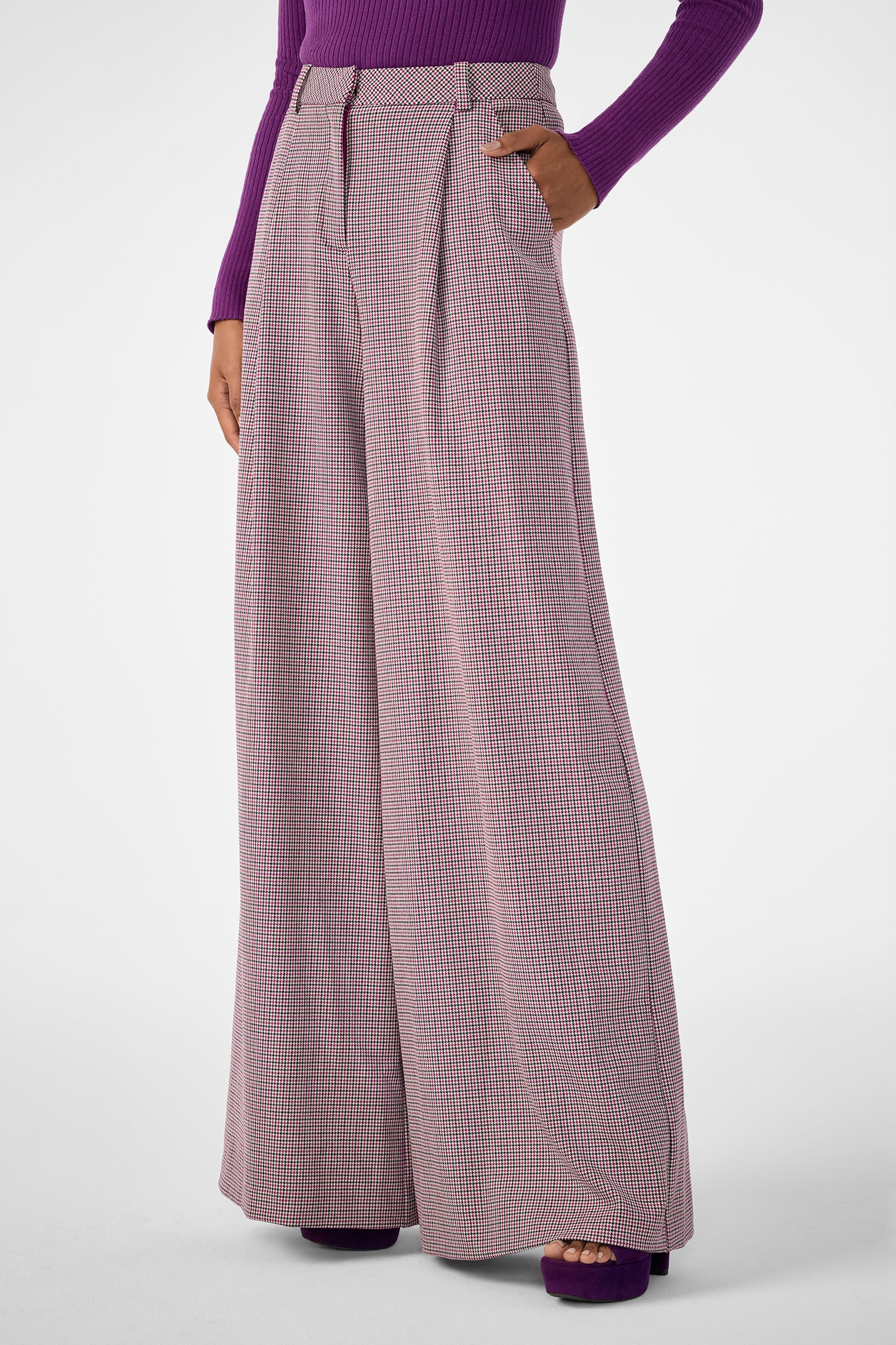 A person in Solani Pant—wide-leg, high-waisted pink and white checkered trousers—hands in pockets, paired with a fitted long-sleeve purple top and matching purple platform shoes.