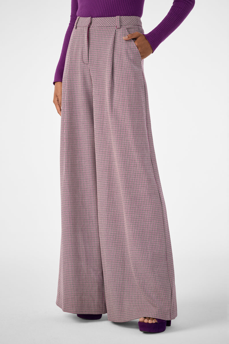 A person in Solani Pant—wide-leg, high-waisted pink and white checkered trousers—hands in pockets, paired with a fitted long-sleeve purple top and matching purple platform shoes.
