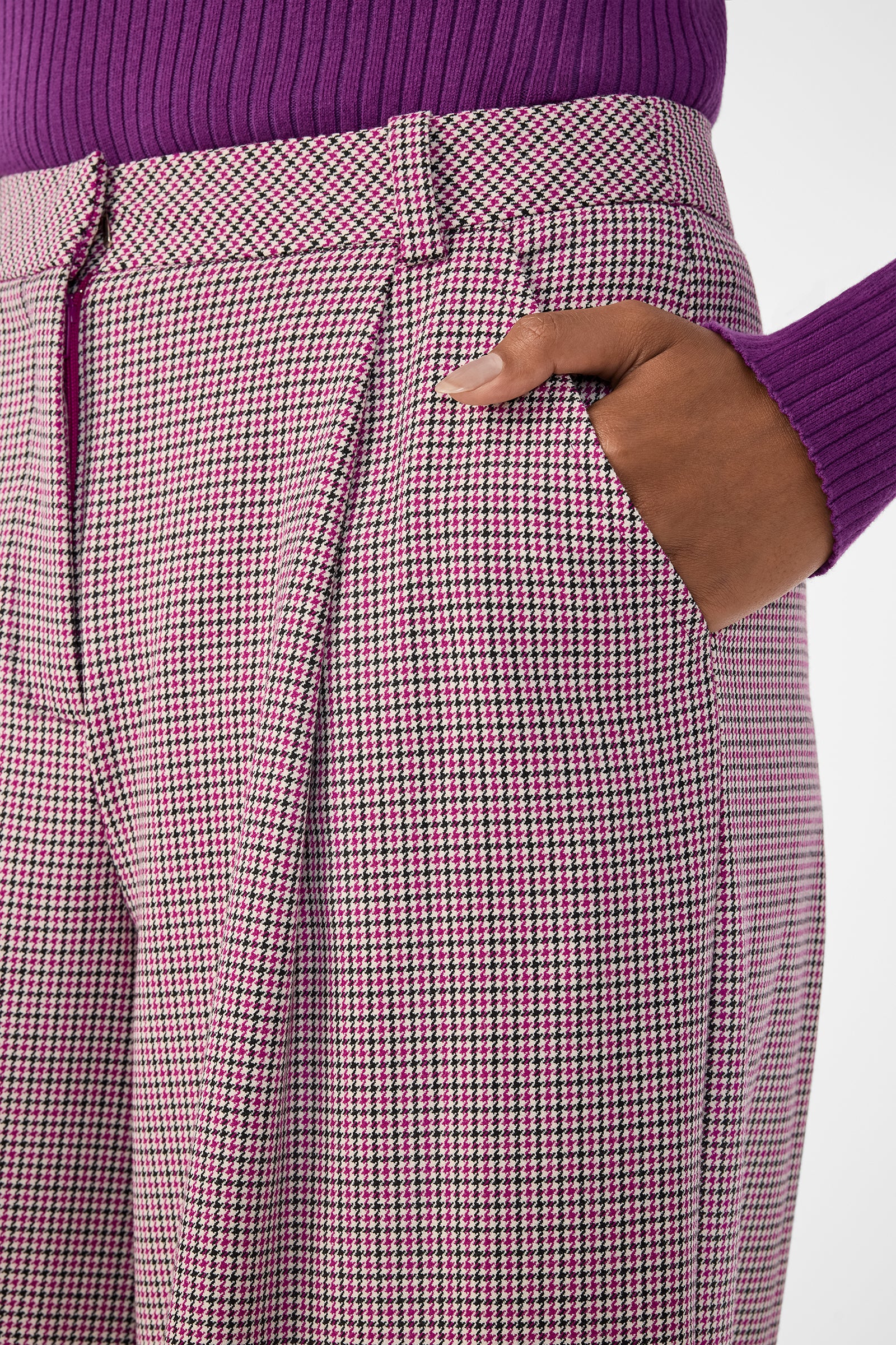 Someone stands with one hand in their pocket, wearing a purple ribbed sweater and high-waisted pink and black Solani Pants.