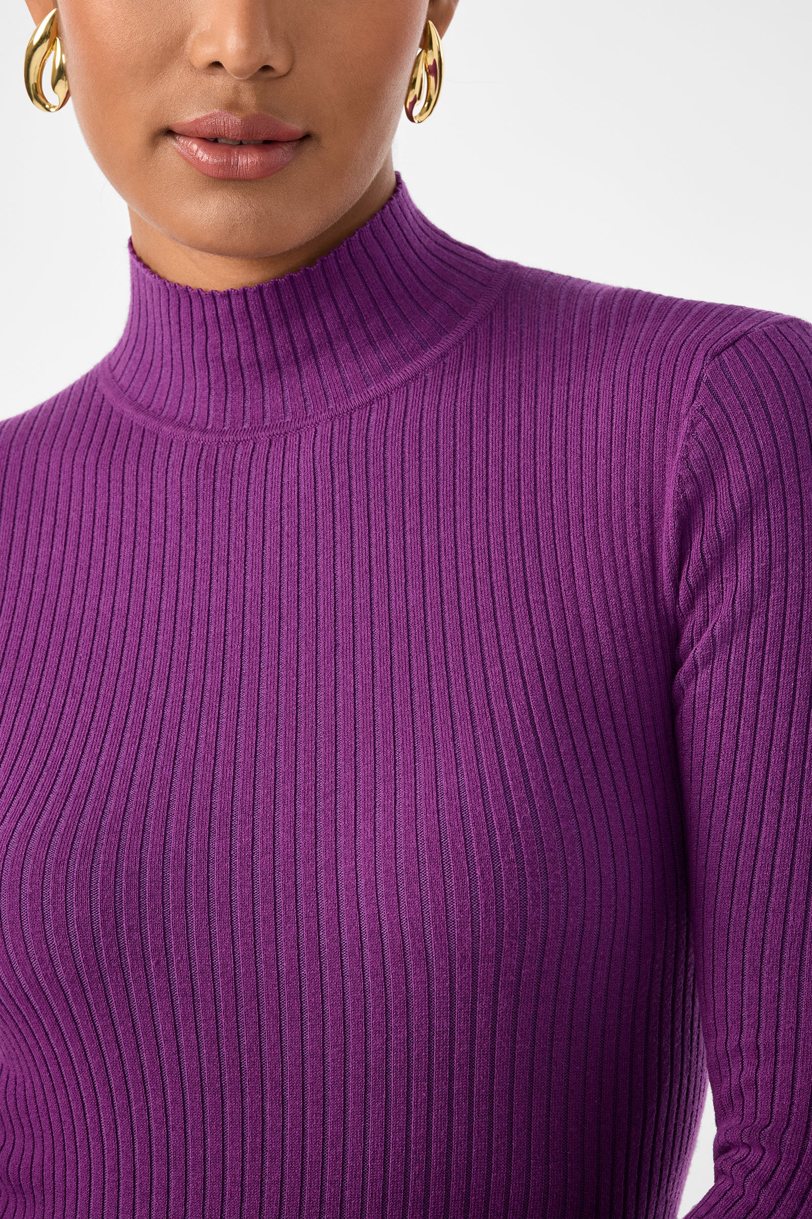A woman wears gold hoop earrings and a purple, ribbed, long-sleeve Hempstead Sweater with a mock neck. Only her lower face, neck, and upper torso appear against a plain white background.