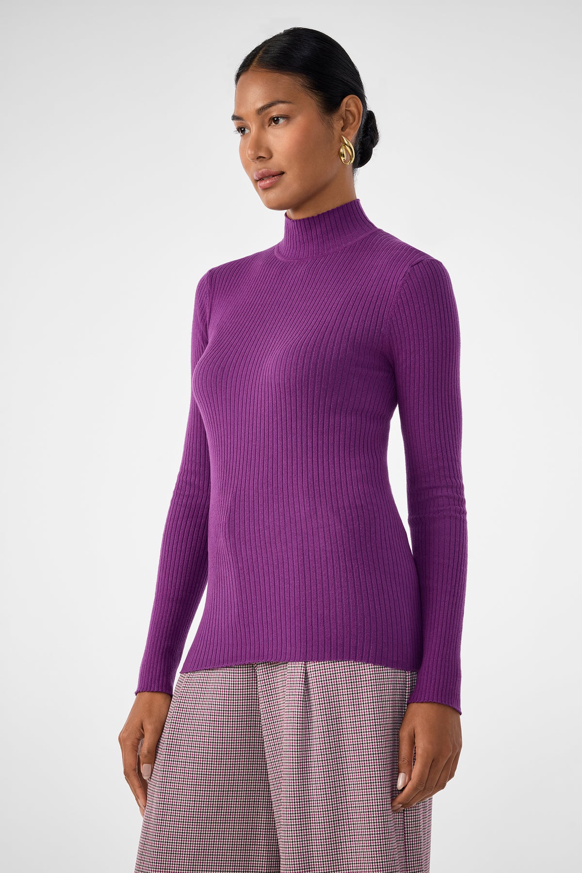 A woman in the fitted, long-sleeve Hempstead Sweater stands against a plain white background, looking slightly to the side while wearing high-waisted, light checkered trousers.