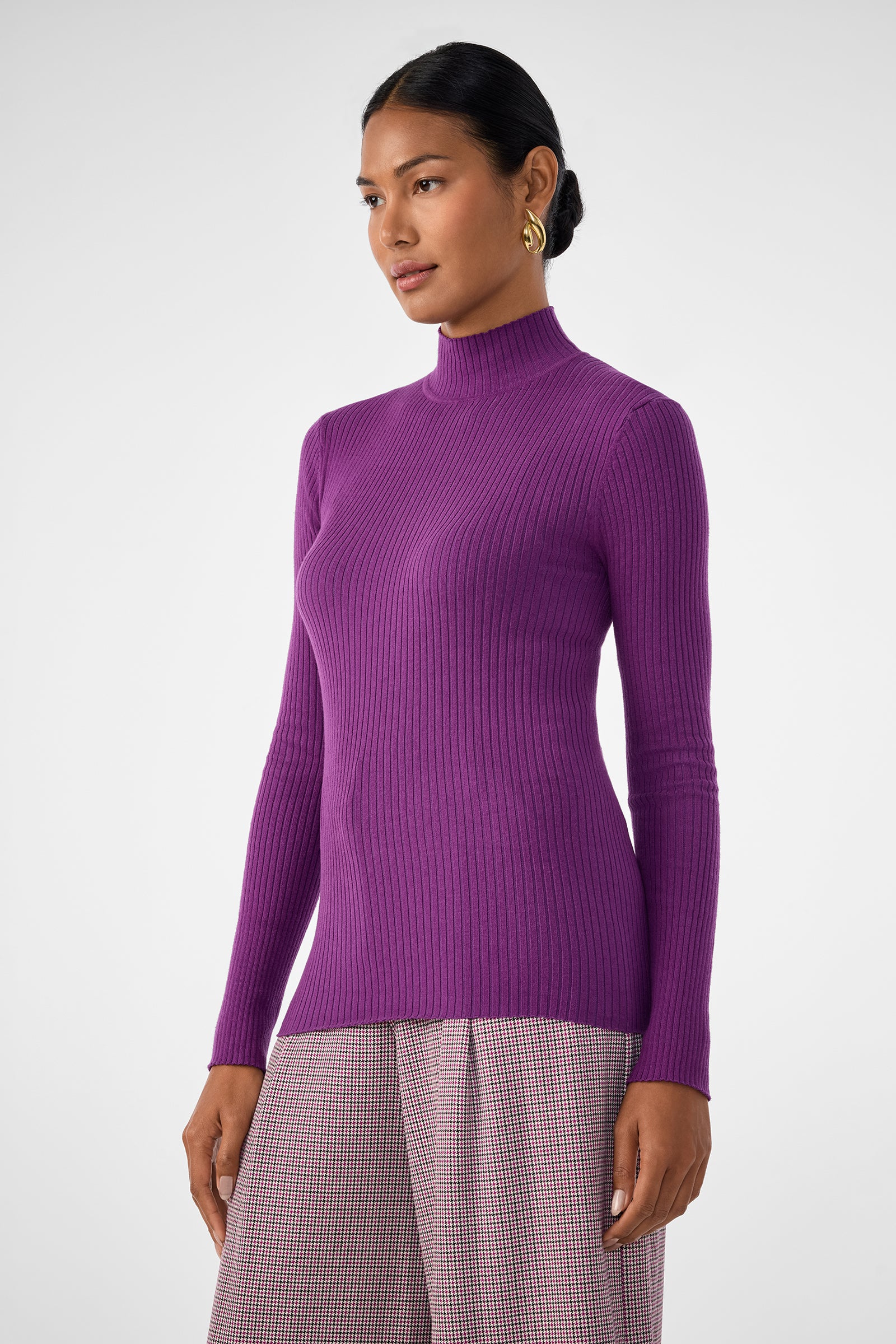 A woman in the fitted, long-sleeve Hempstead Sweater stands against a plain white background, looking slightly to the side while wearing high-waisted, light checkered trousers.