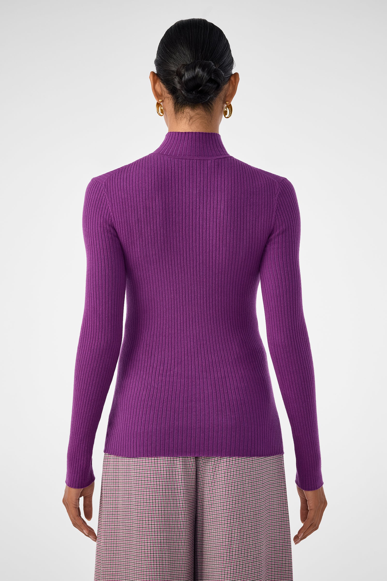 A person stands facing away, wearing the fitted, long-sleeve Hempstead Sweater in purple with a high-waisted checkered skirt. Their hair is in a neat bun and they accessorize with gold hoop earrings.