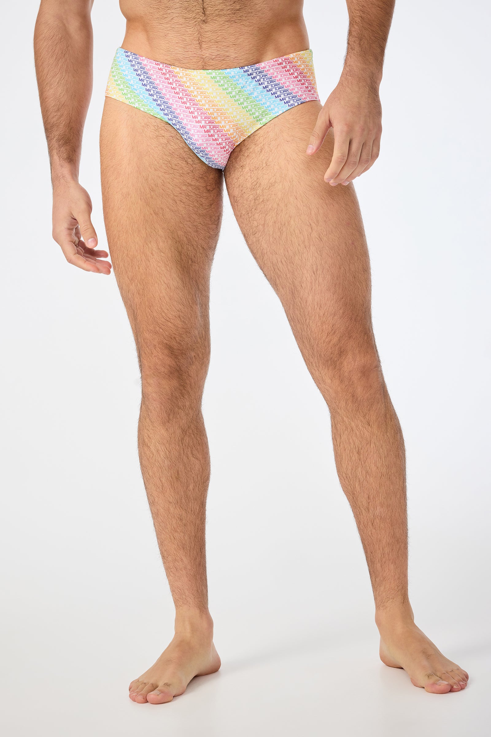 Model wears the Bondi Swim Brief - Pride in multi, standing barefoot against a white background with arms relaxed by their sides; only their body from shoulders to feet is visible.