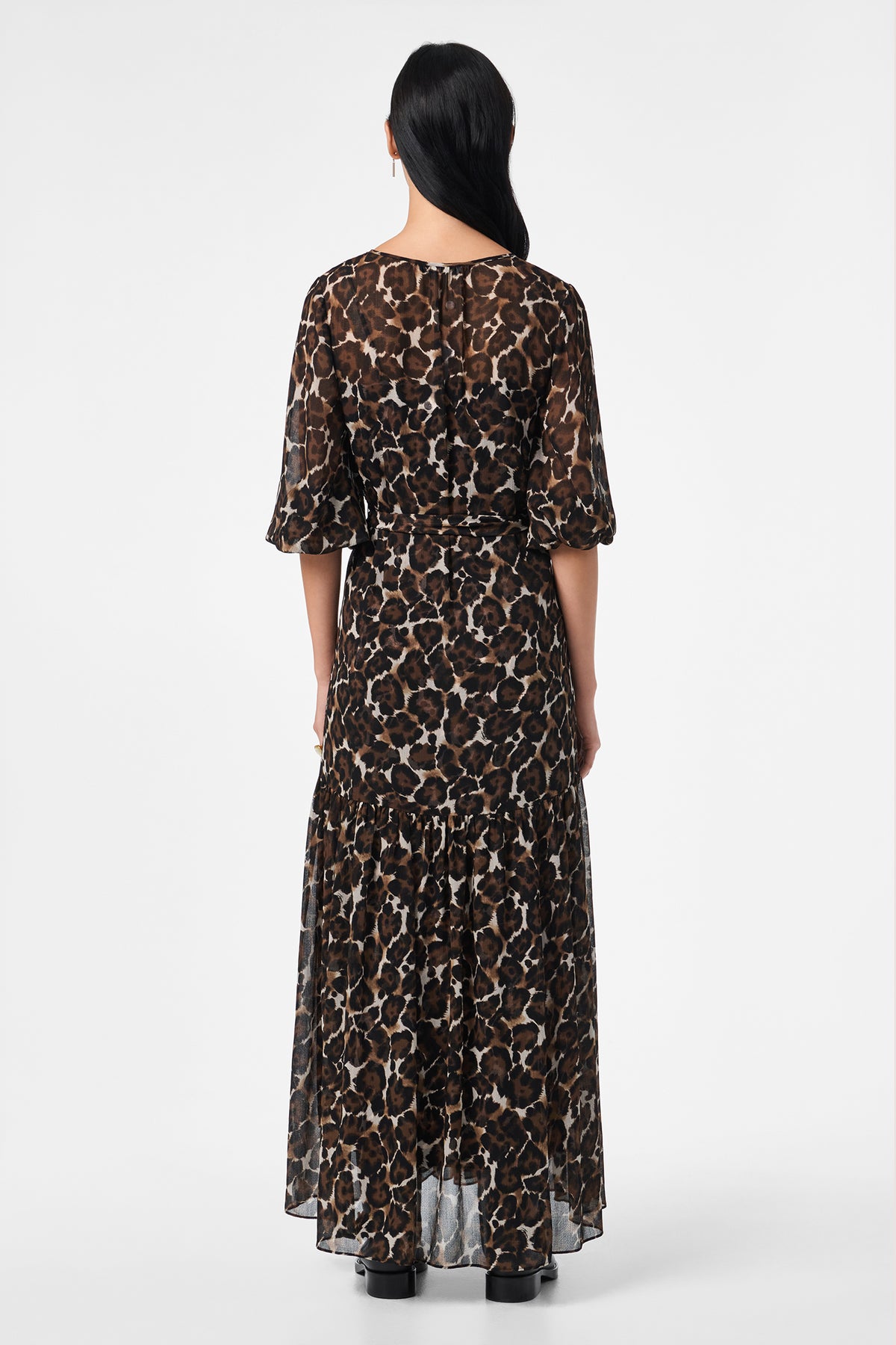 A person with long dark hair faces away, wearing the Shalina Tie-Waist Maxi Dress featuring a brown and black animal print, ankle length, and elbow-length sleeves against a plain white background.