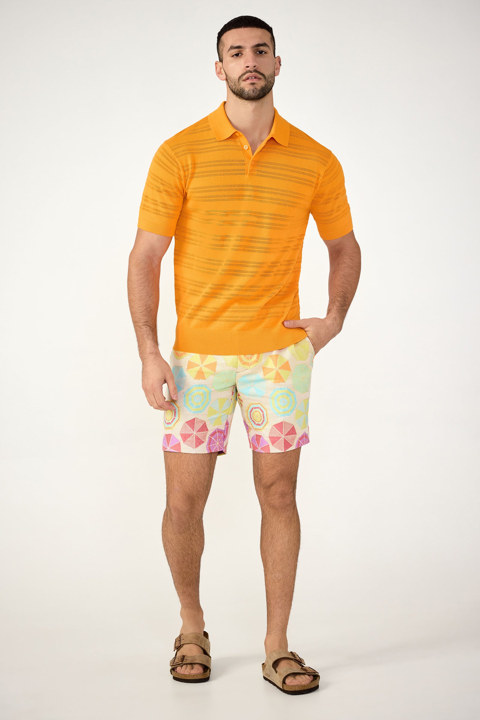 A man stands against a plain background wearing the Edison Short-Sleeve Striped Polo, pastel patterned shorts, and brown sandals, with one hand in his pocket and a neutral expression.