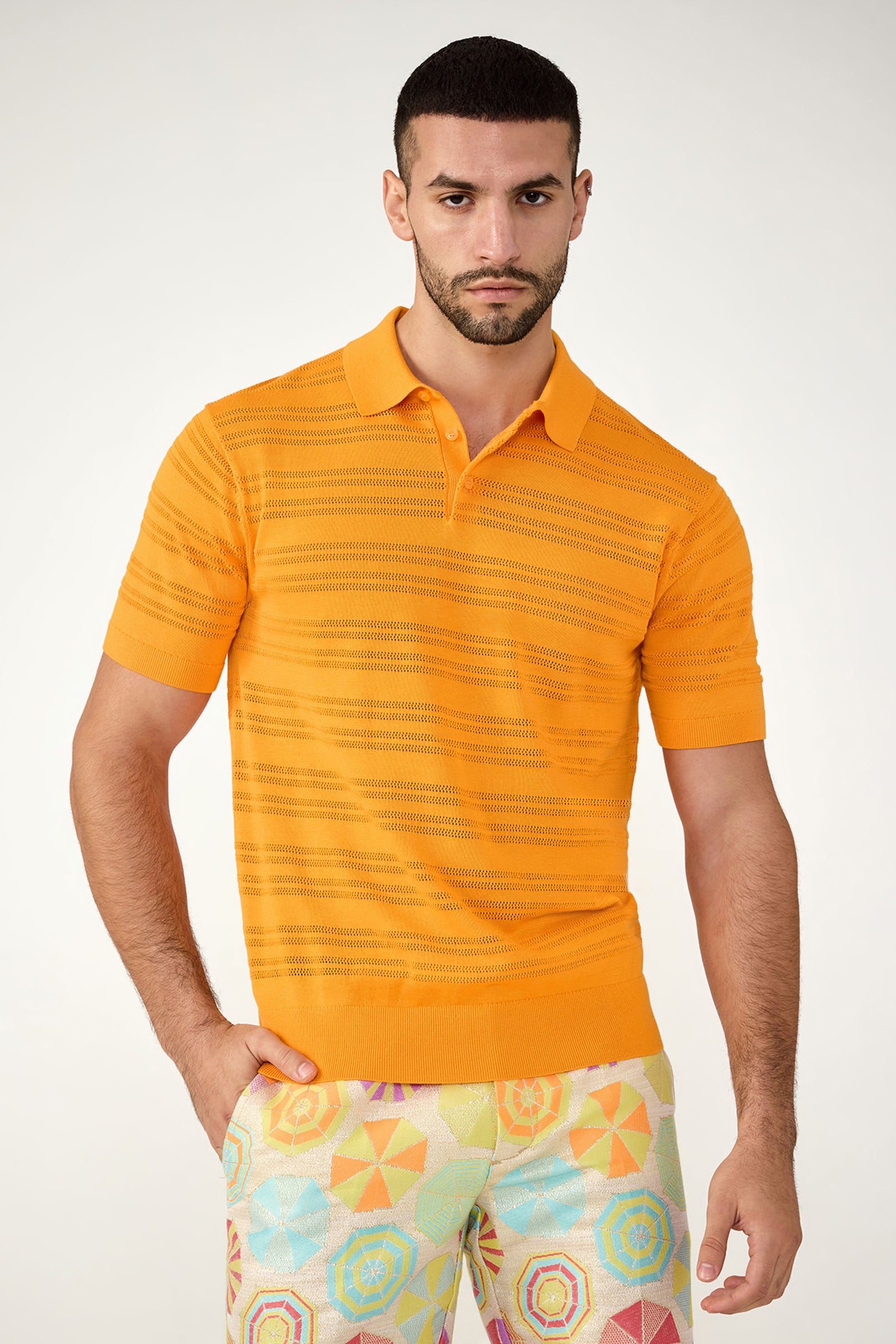 A man with short dark hair and a beard wears the Edison Short-Sleeve Striped Polo in Cap Clementine, paired with colorful geometric-patterned pants, standing against a plain light background.
