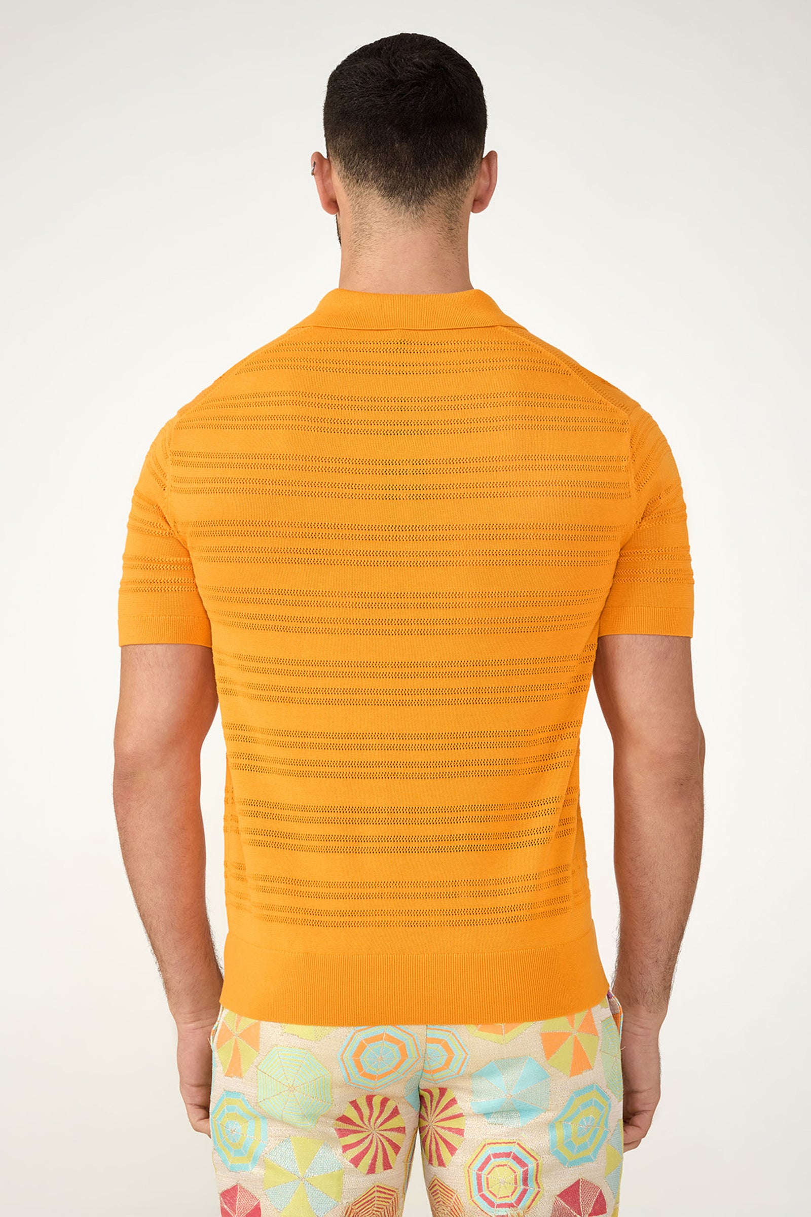 A man is seen from behind wearing the Edison Short-Sleeve Striped Polo in bright orange, paired with multicolored geometric shorts. The background is plain and light-colored.