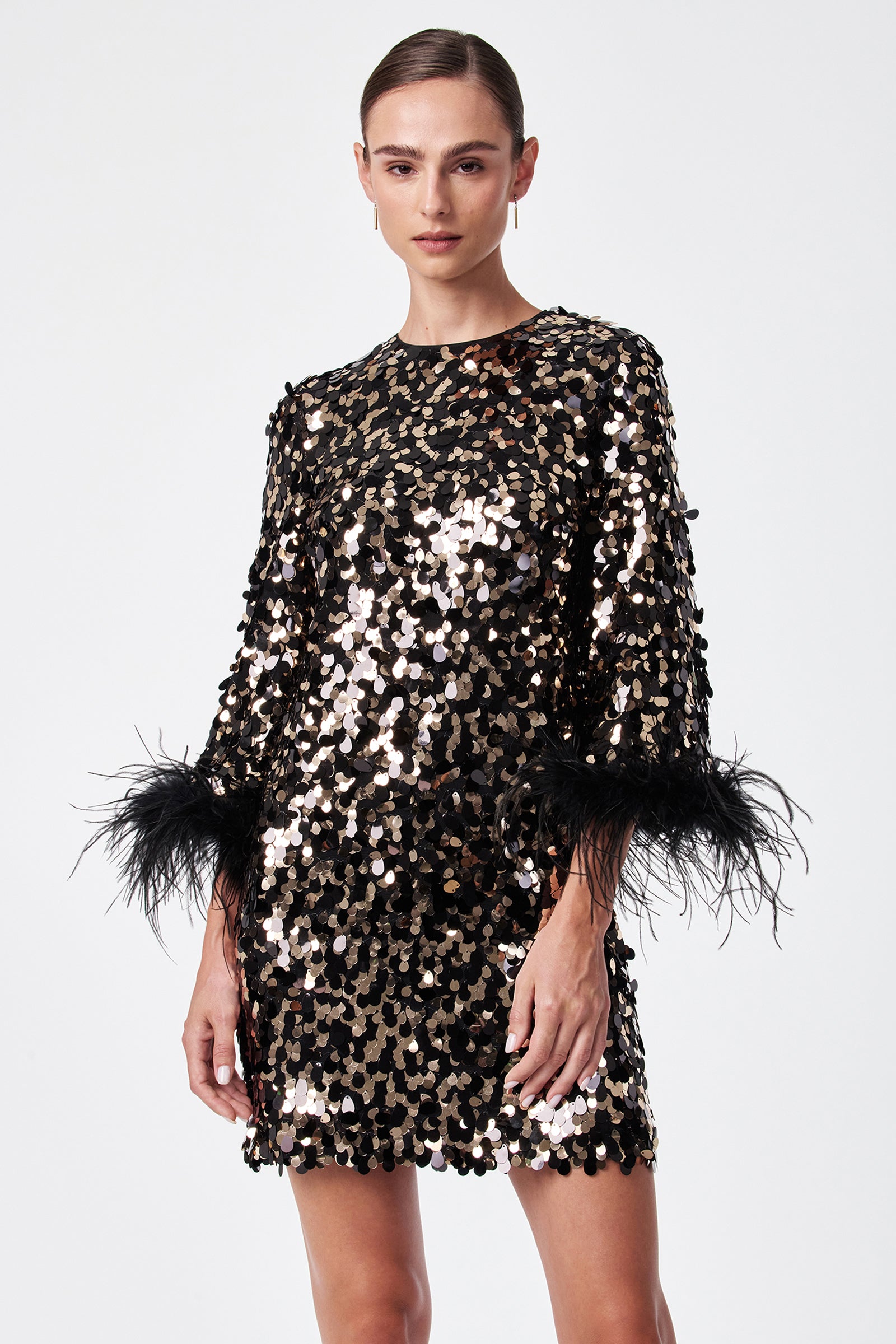 A woman models the Juliet Dress, a black and gold sequin outfit with black feather-accented sleeves, standing against a plain white background.