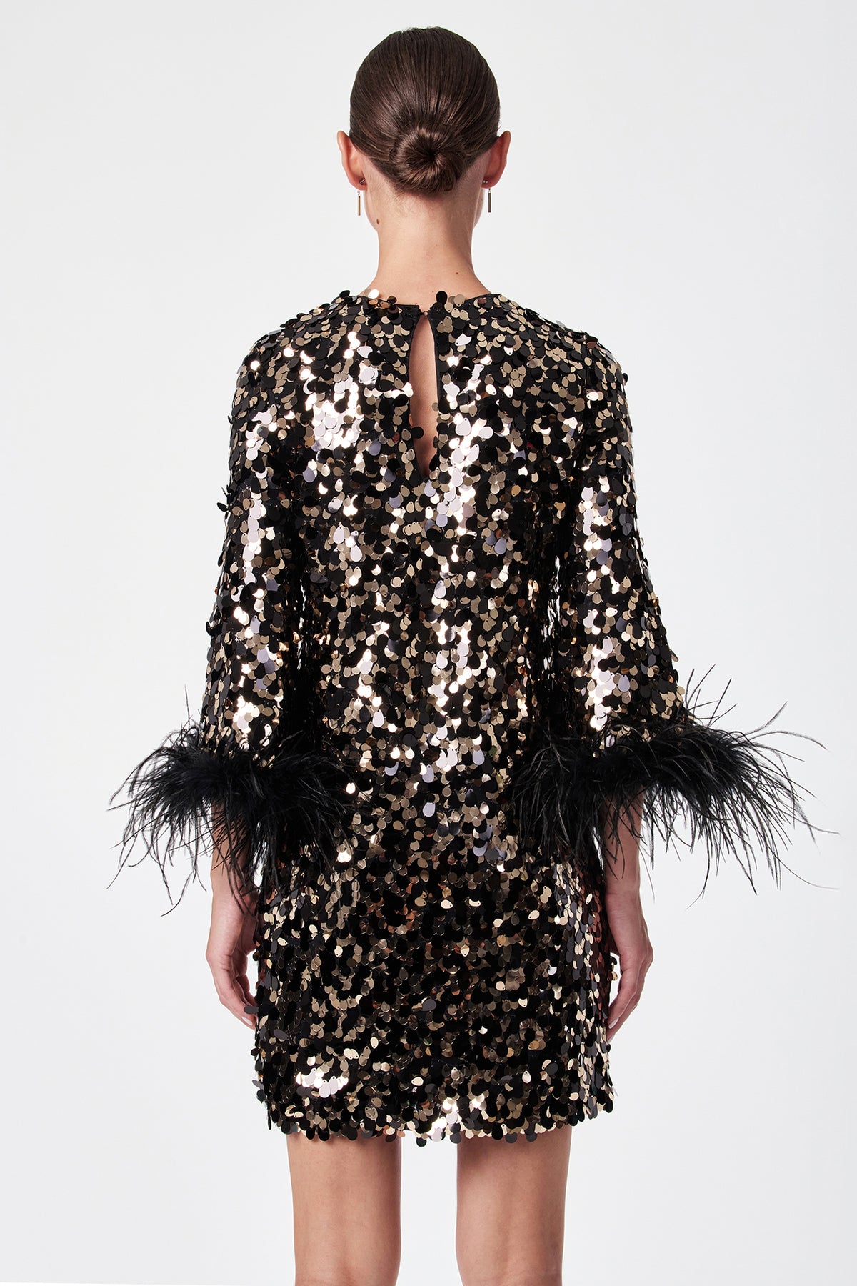 A woman faces away, wearing the Juliet Dress—a short black dress adorned with gold and silver sequins and black feather sleeve trim. Her hair is in a sleek bun, and she stands against a plain white background.