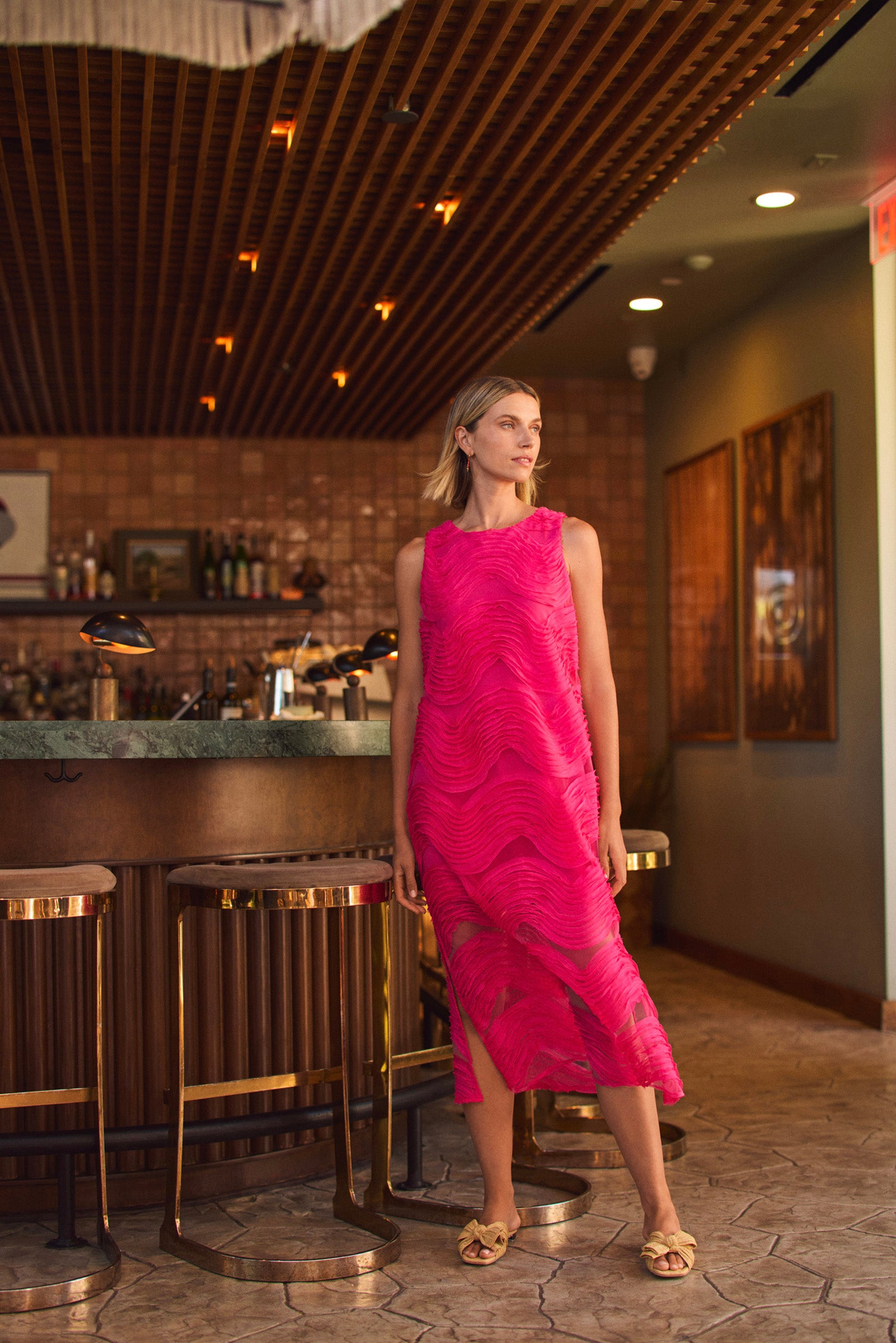 A woman in the vibrant pink Belisama Organza Midi Dress stands near a bar with gold barstools, surrounded by a stylish, warmly lit interior featuring wood and marble accents.