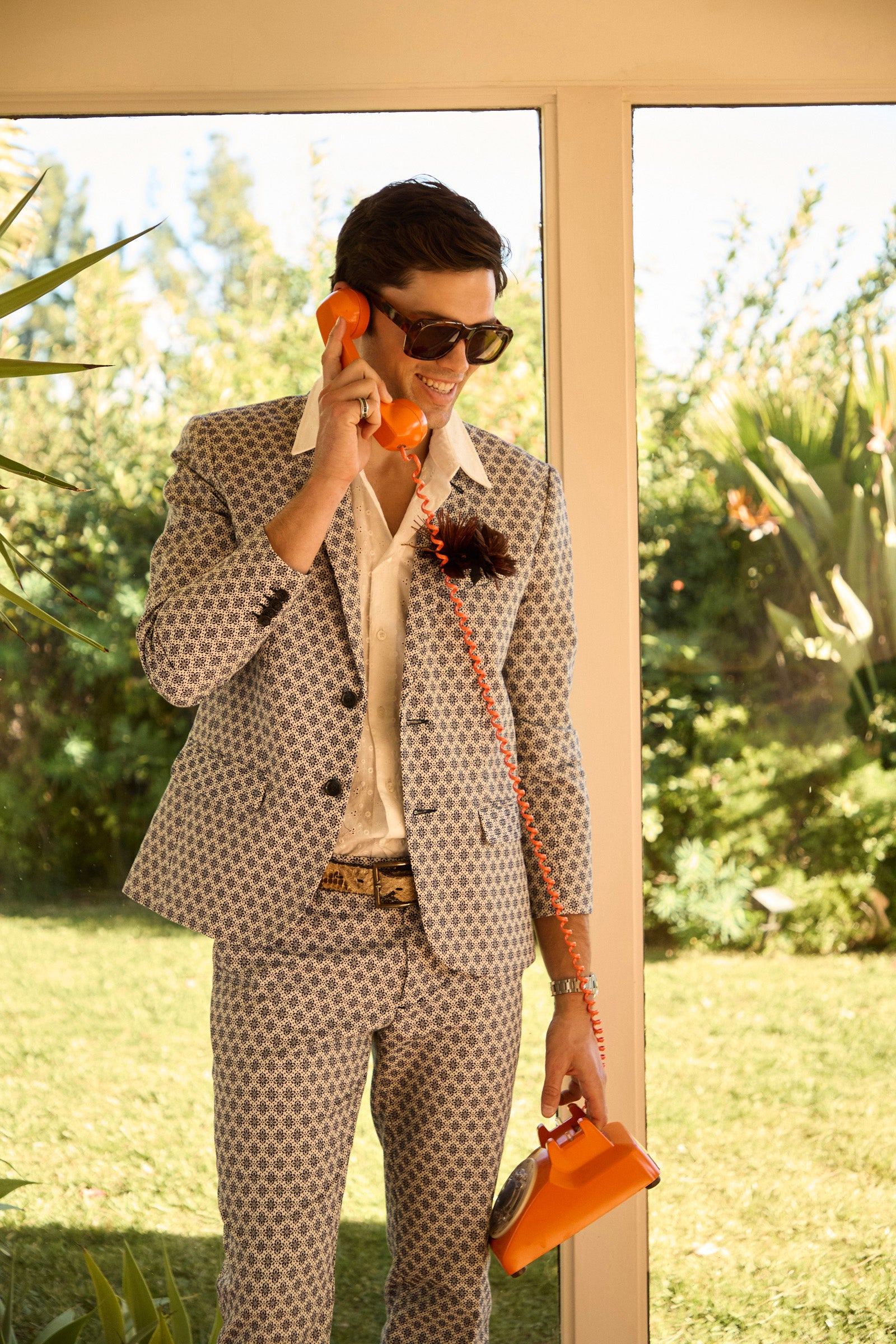 A man wearing the Thurston Blazer - Daisy Jacquard and sunglasses smiles as he talks on an orange rotary phone, standing indoors by a glass wall with greenery and sunlight outside.